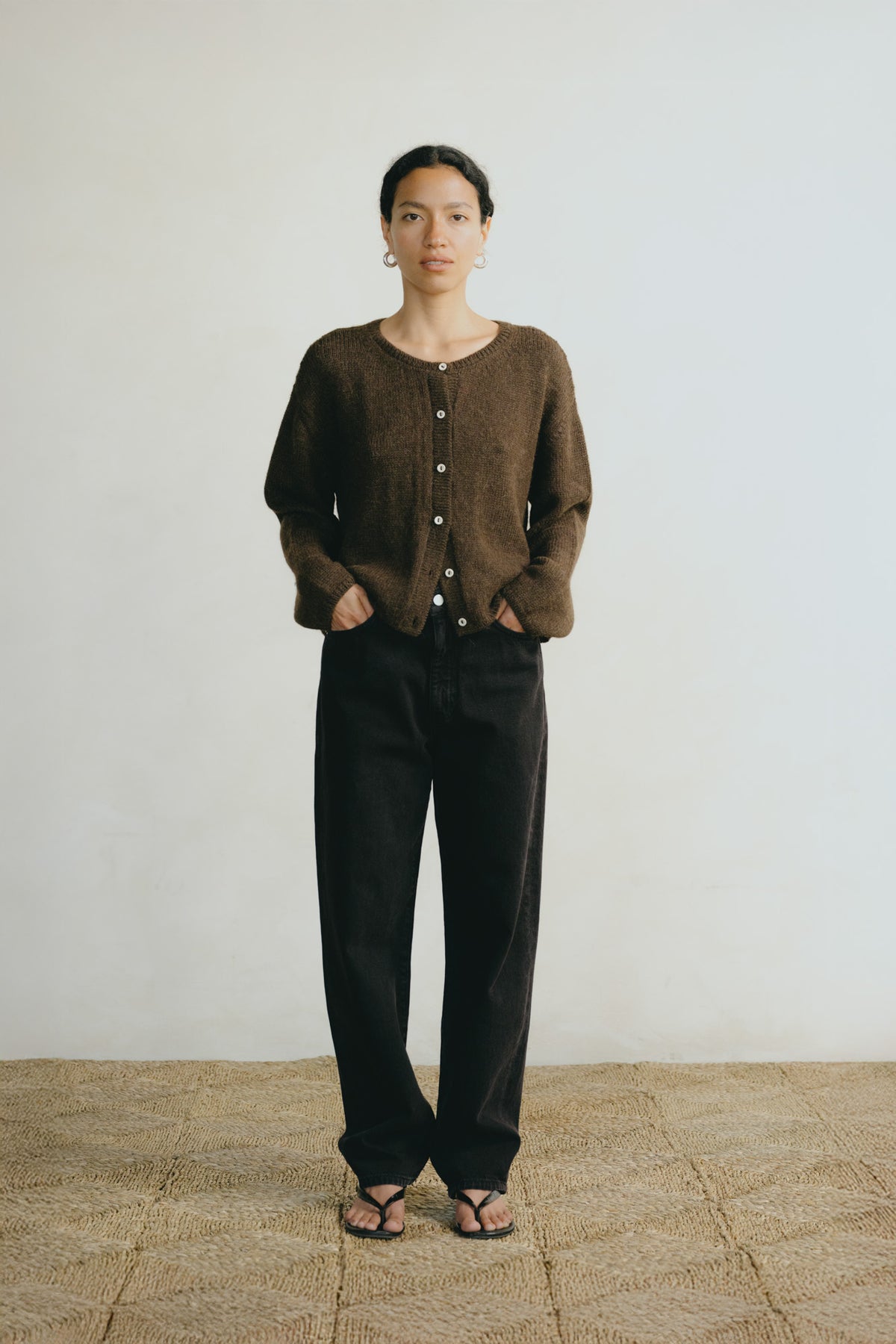 A model stands against a plain off-white wall wearing a brown button-front cardigan, loose black pants, and sandals.