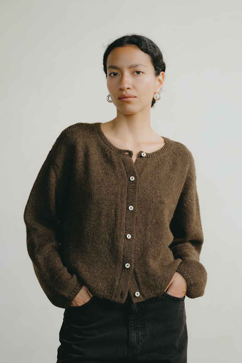 A woman wearing a brown knit button-front cardigan sweater and black pants, standing against a plain light gray background.