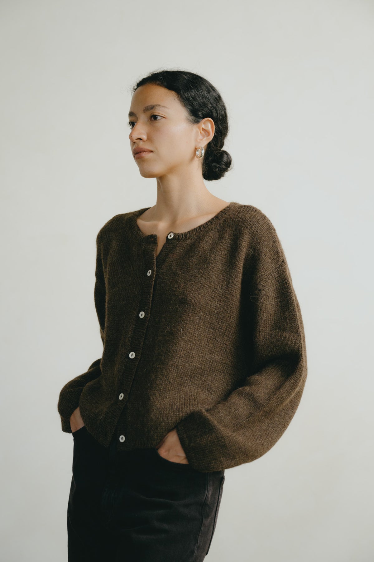 A woman wearing a dark brown fuzzy button-up cardigan sweater and black pants, standing against a plain light gray background.