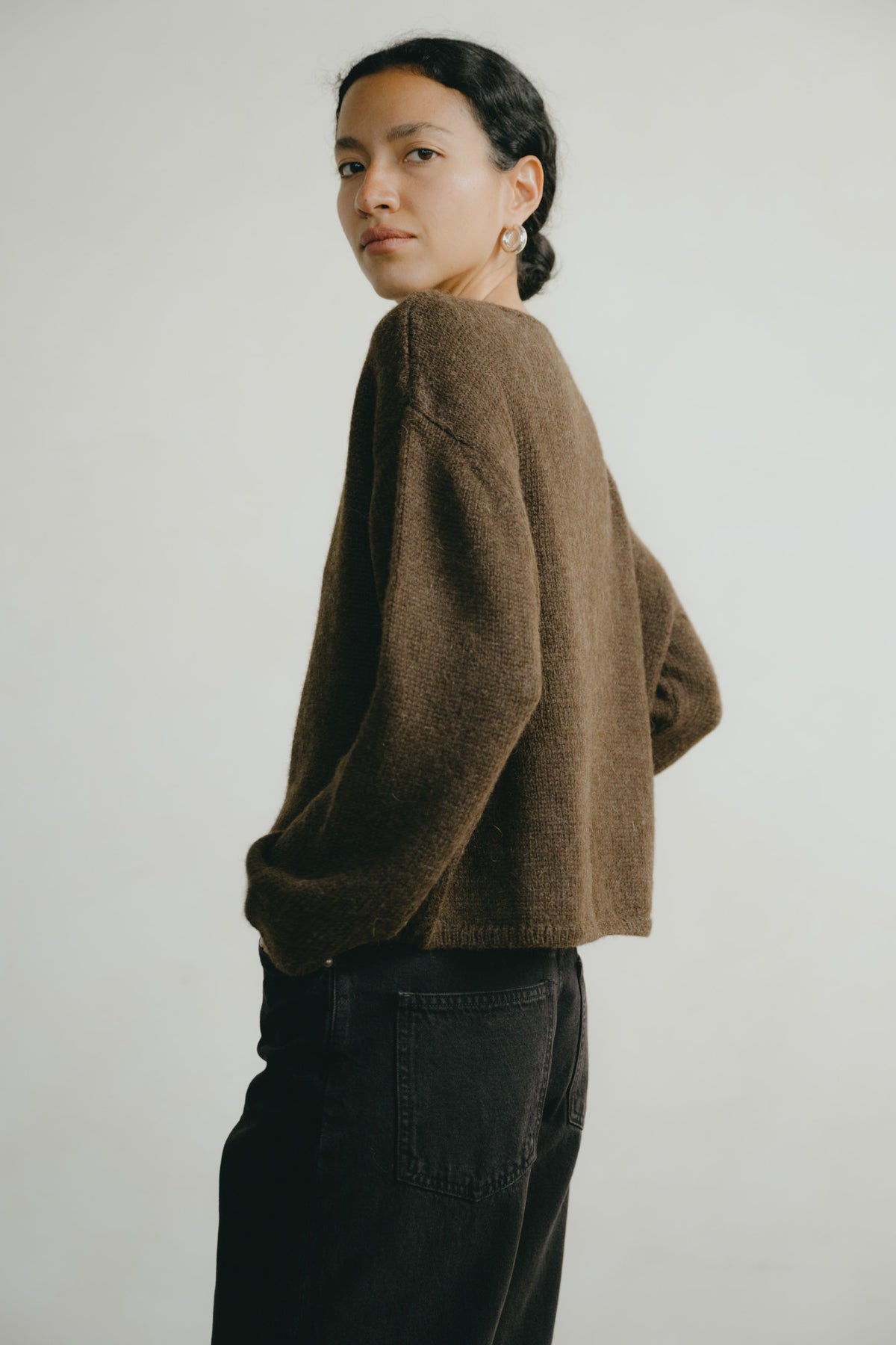 A woman wearing a brown fuzzy cropped sweater and dark jeans, photographed from the back against a plain light gray studio background.