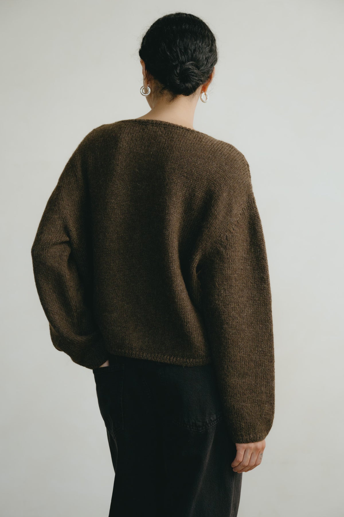 A person shown from the back wearing a loose, textured brown knit sweater and dark pants against a plain light gray background.