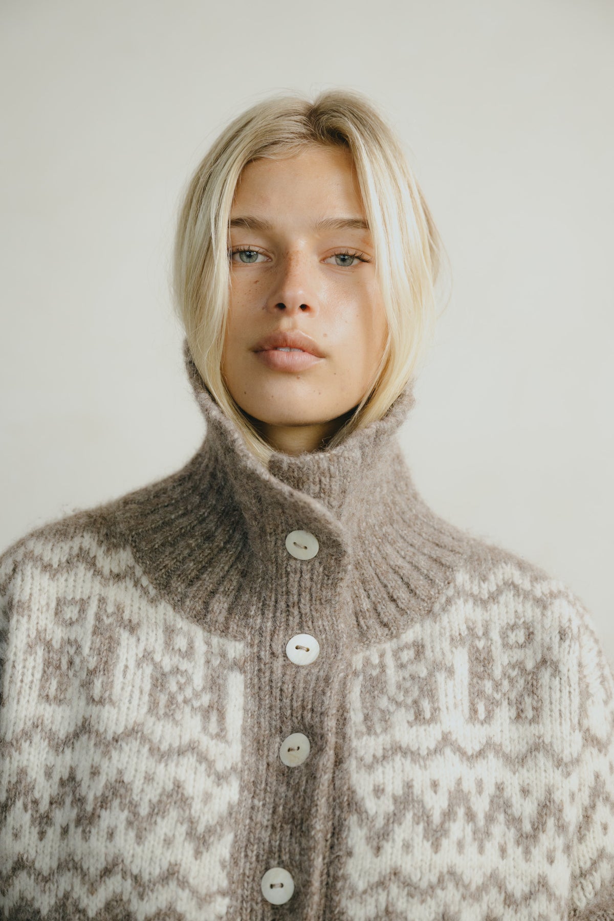 A close-up portrait of a blonde woman wearing a high-neck, button-front knit sweater with a brown and cream Fair Isle pattern, set against a plain light background.