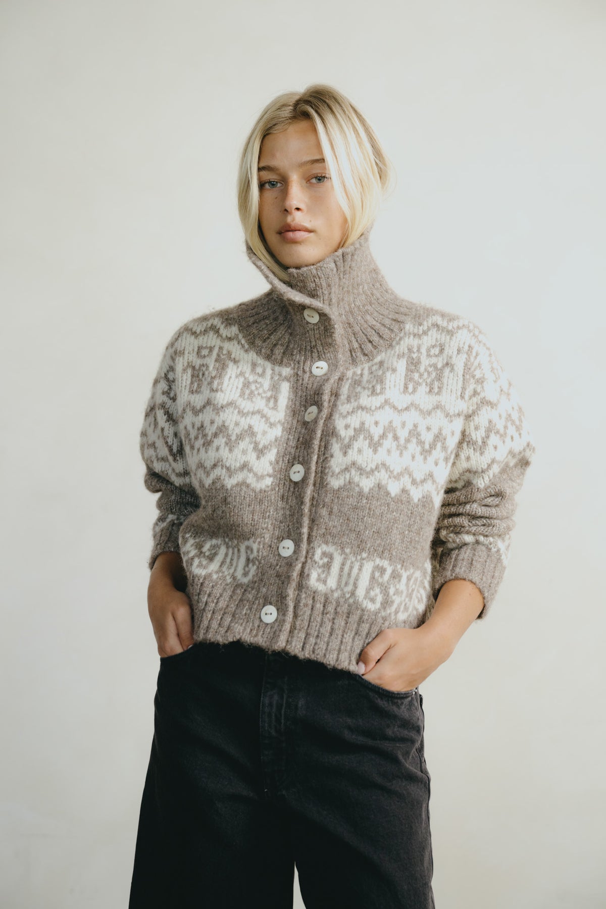 A blonde woman wearing a cropped, high-neck knit cardigan with a brown and cream Fair Isle pattern, paired with black jeans, against a plain light gray background.