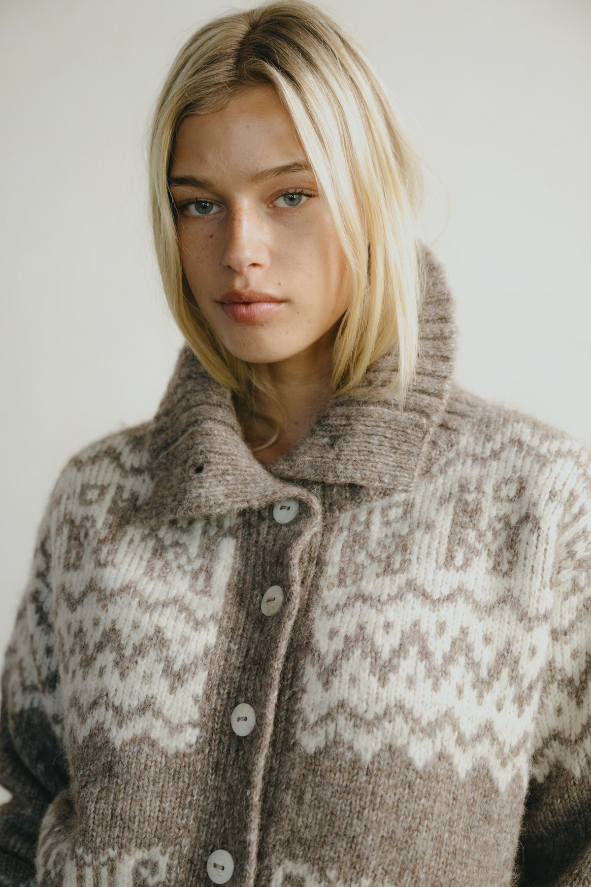 A close-up of a blonde woman wearing a brown and cream patterned knit cardigan sweater with a high collar and front buttons, set against a plain light background.