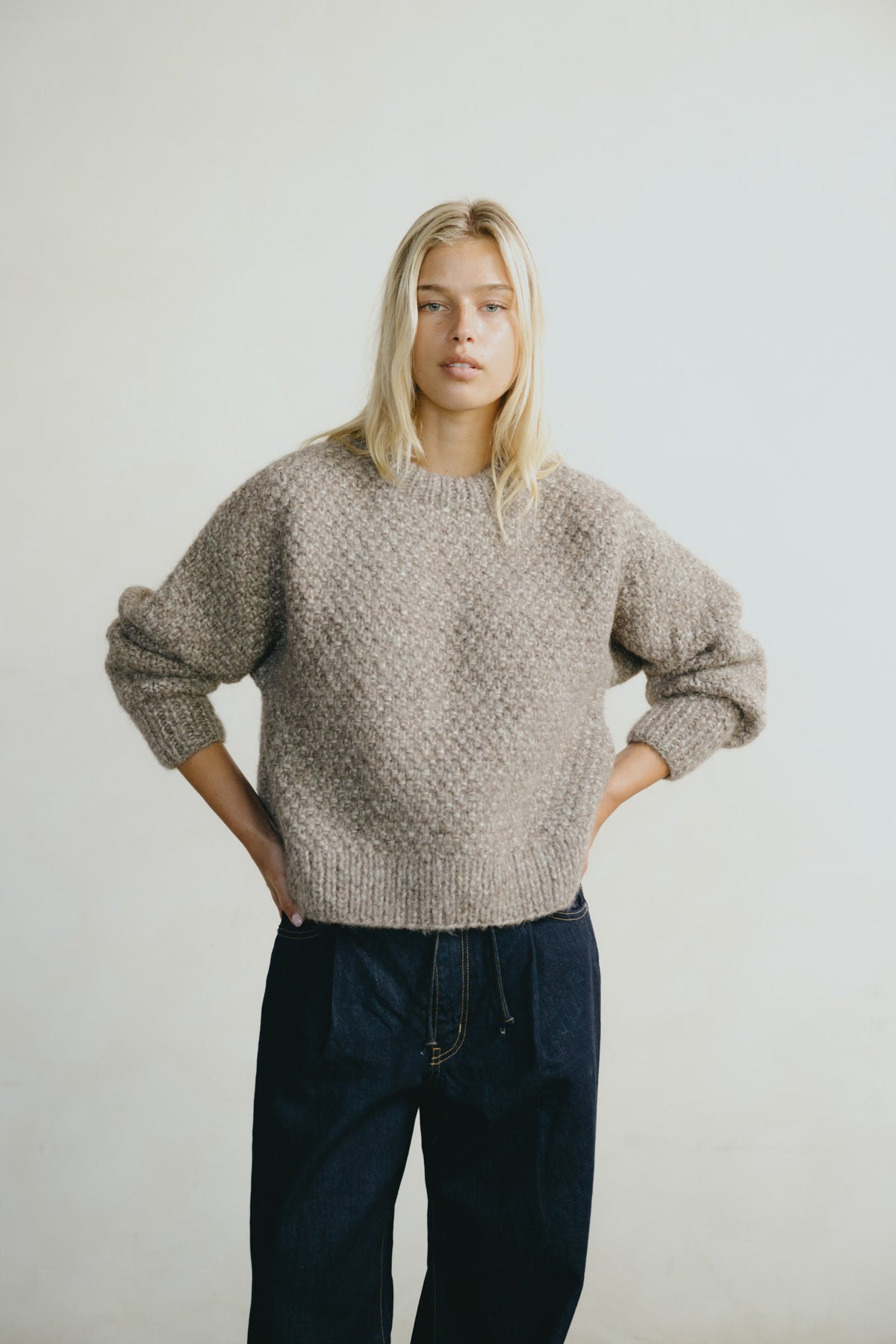 A blonde woman wearing a chunky, textured knit crewneck sweater in a taupe-beige color, paired with dark blue jeans, standing against a plain light gray studio background.