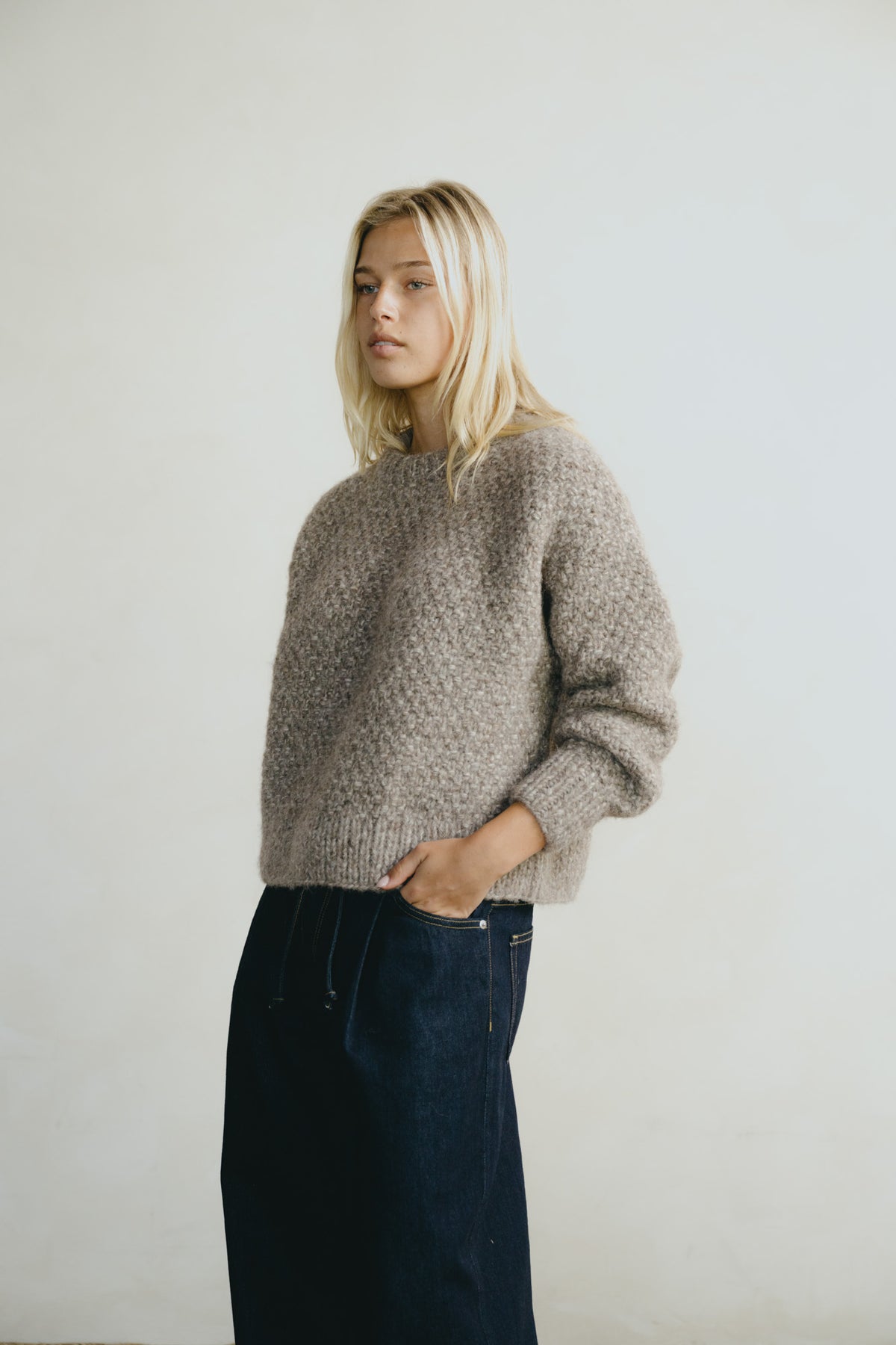 A blonde woman wearing a chunky, textured taupe knit sweater and dark blue jeans, standing against a plain off-white wall.