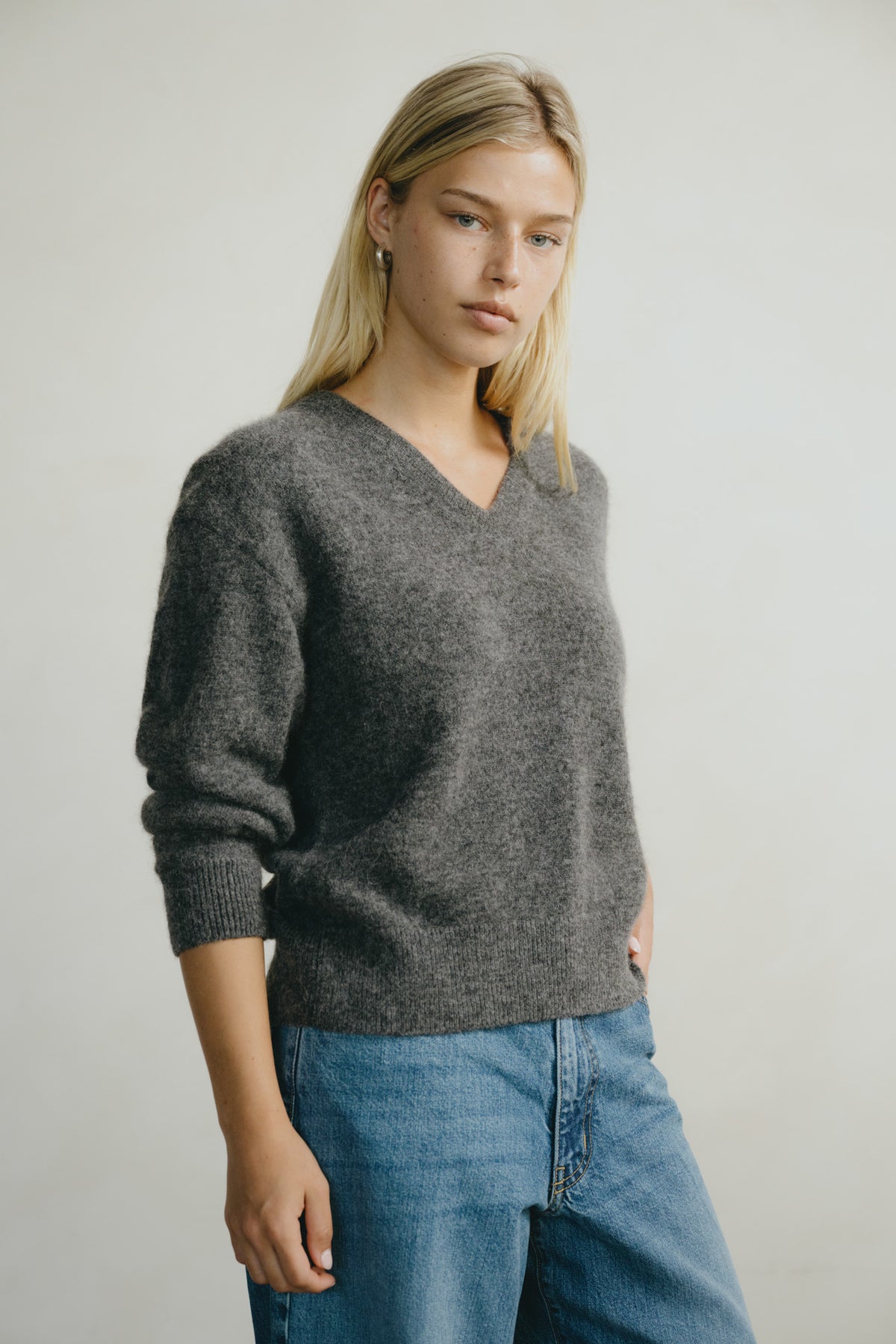 A woman wearing a dark gray heathered V-neck knit sweater and blue jeans, standing against a plain light gray studio background.