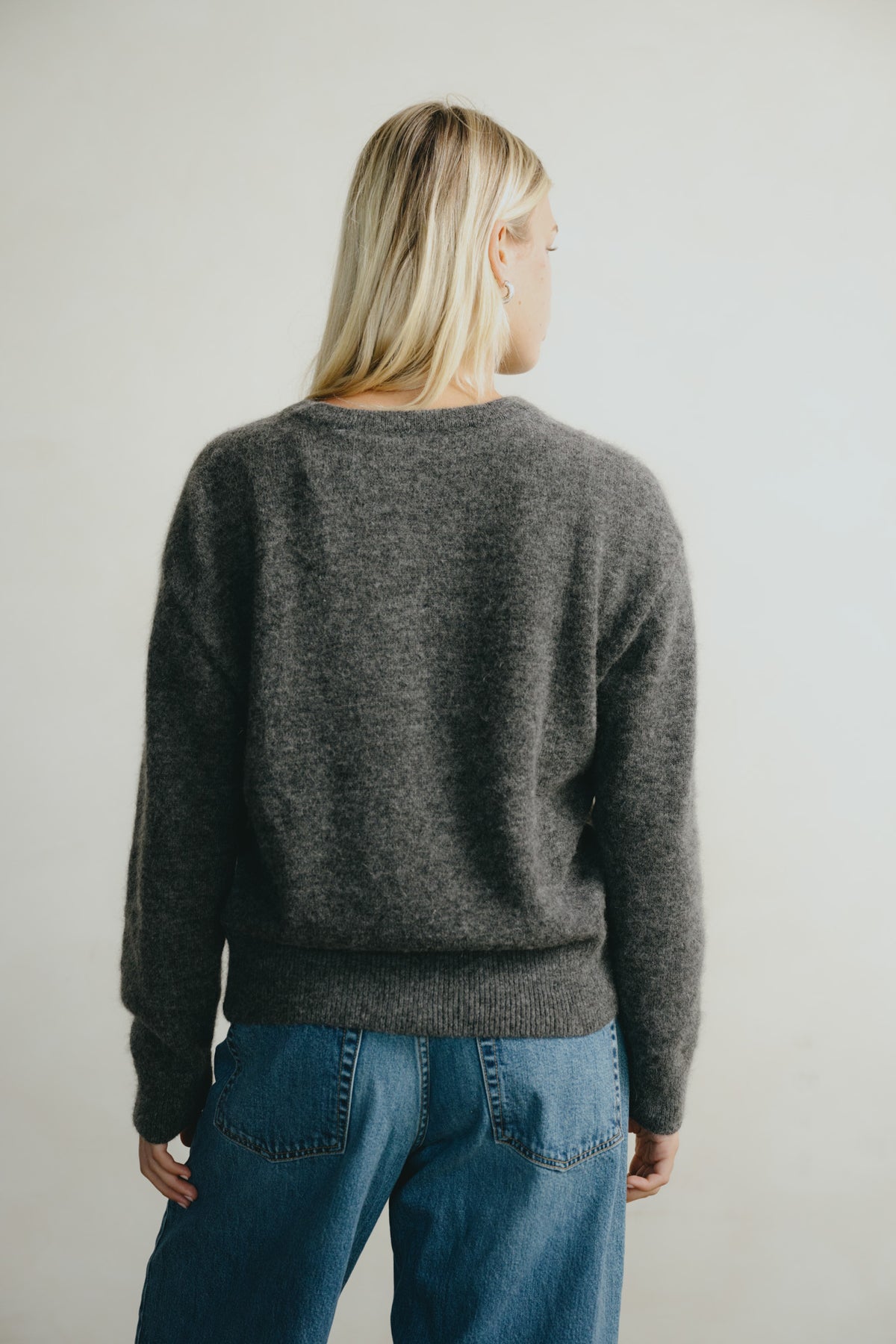 A woman with blonde hair is shown from behind wearing a dark gray heathered knit sweater and blue jeans against a plain light gray background.