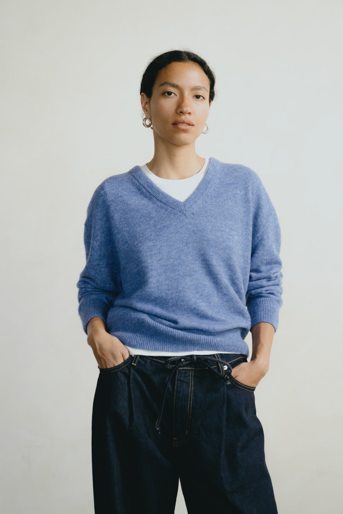 A model wearing a light blue v-neck knit sweater over a white undershirt and dark blue jeans, standing against a plain light gray background.