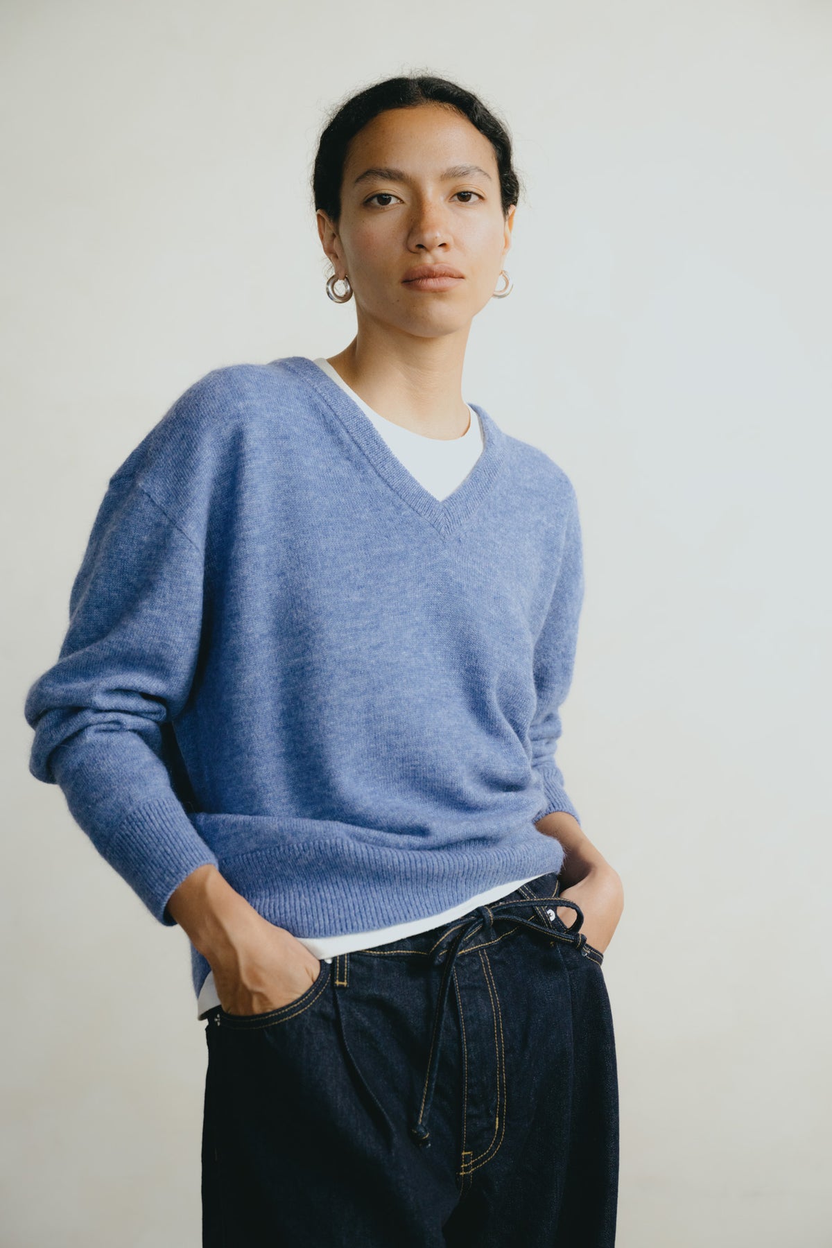 A model wearing a heathered periwinkle blue V-neck knit sweater over a white undershirt and dark blue jeans, standing against a plain off-white background.