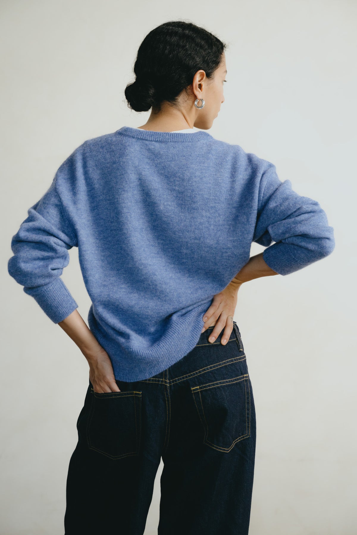 A woman shown from the back wearing a heathered blue knit sweater and dark denim jeans against a plain light gray background.