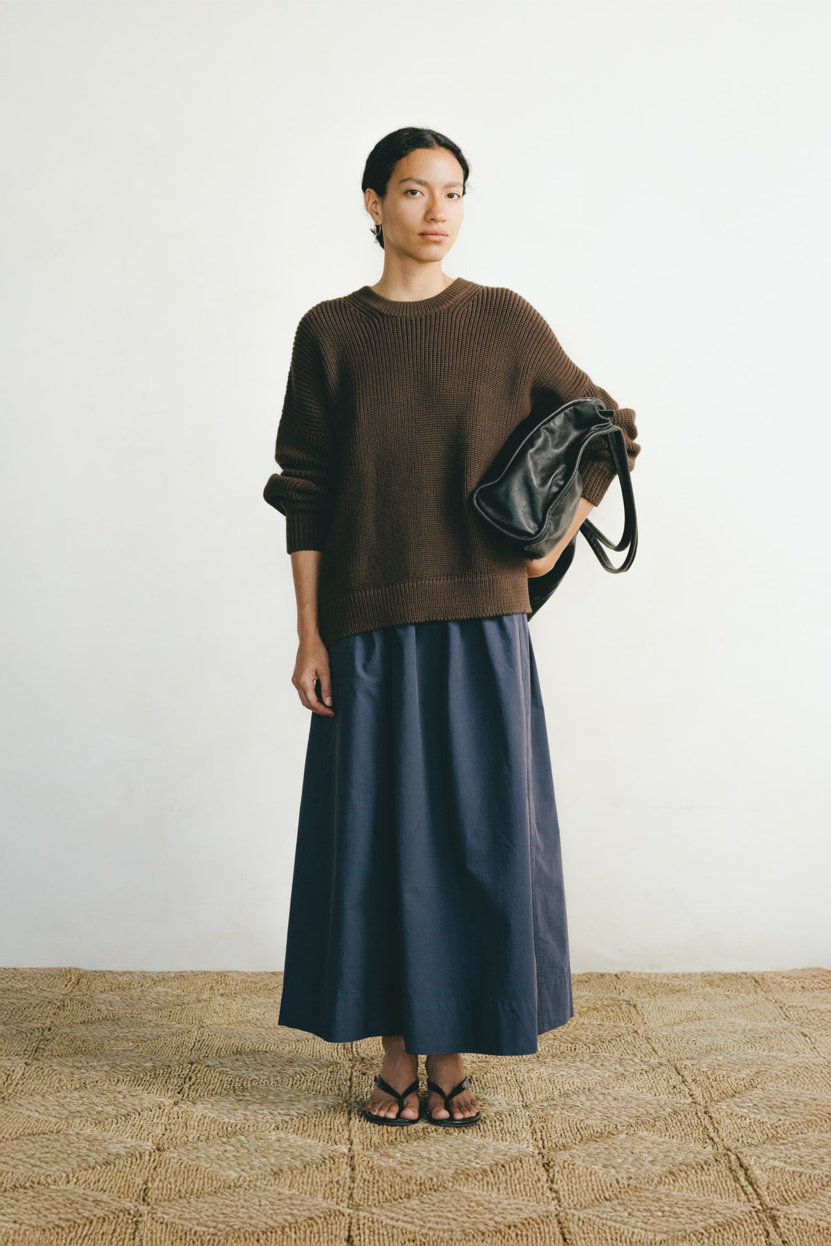 A model stands against a plain off-white wall wearing a brown knit crewneck sweater and a long navy blue skirt, holding a black shoulder bag.