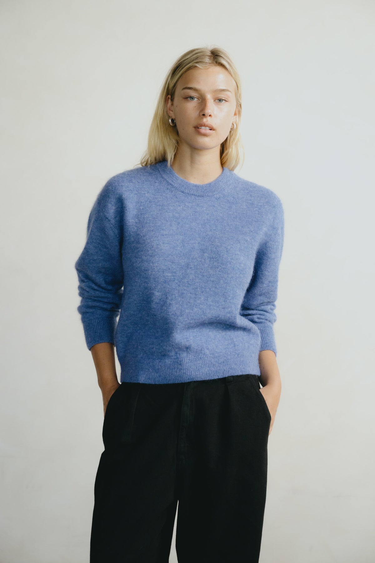 A blonde woman wearing a heathered periwinkle blue crewneck sweater and black trousers, standing against a plain light gray background.