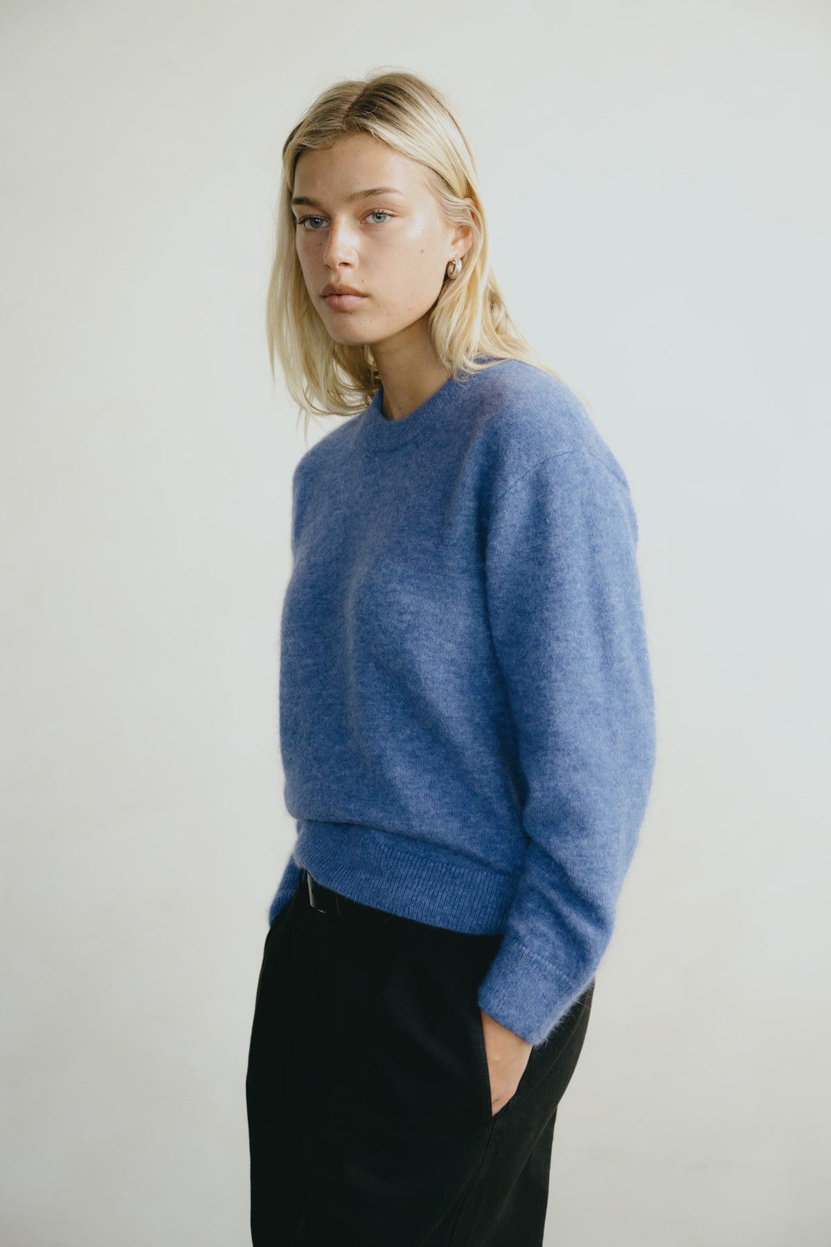 A blonde woman wearing a soft heathered blue crewneck sweater and black trousers, standing against a plain light gray studio background.