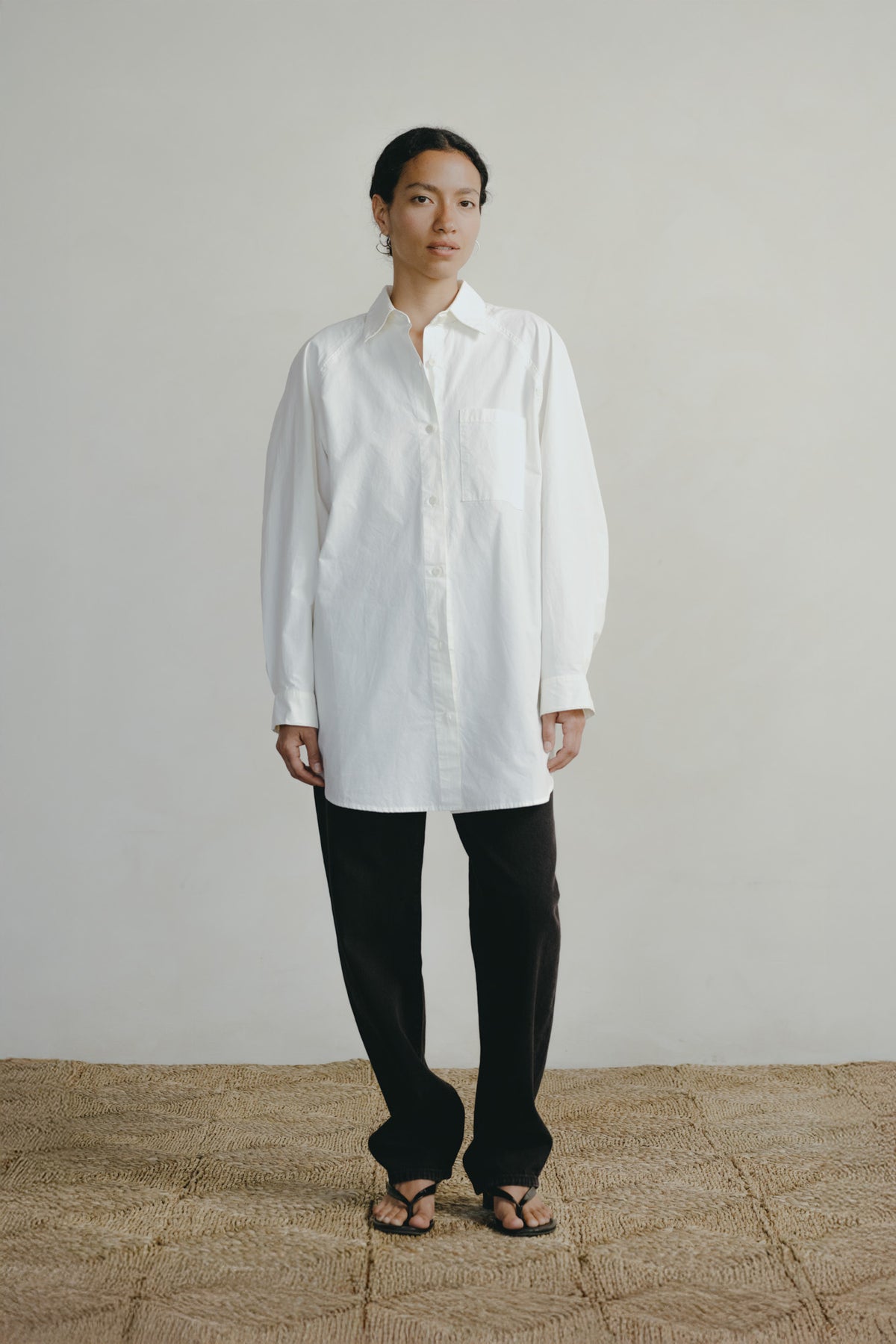 A model stands against a light beige wall and textured tan floor, wearing a loose white long-sleeve button-up shirt, black wide-leg pants, and black sandals.