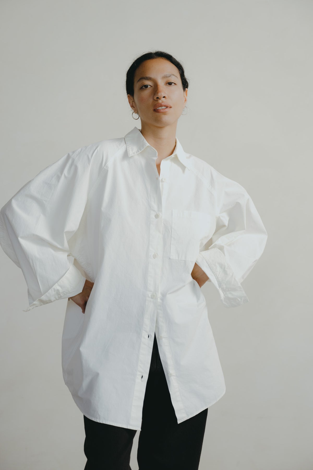 A model wearing an oversized white button-down shirt with dramatic balloon sleeves and black pants, standing against a plain light gray studio background.