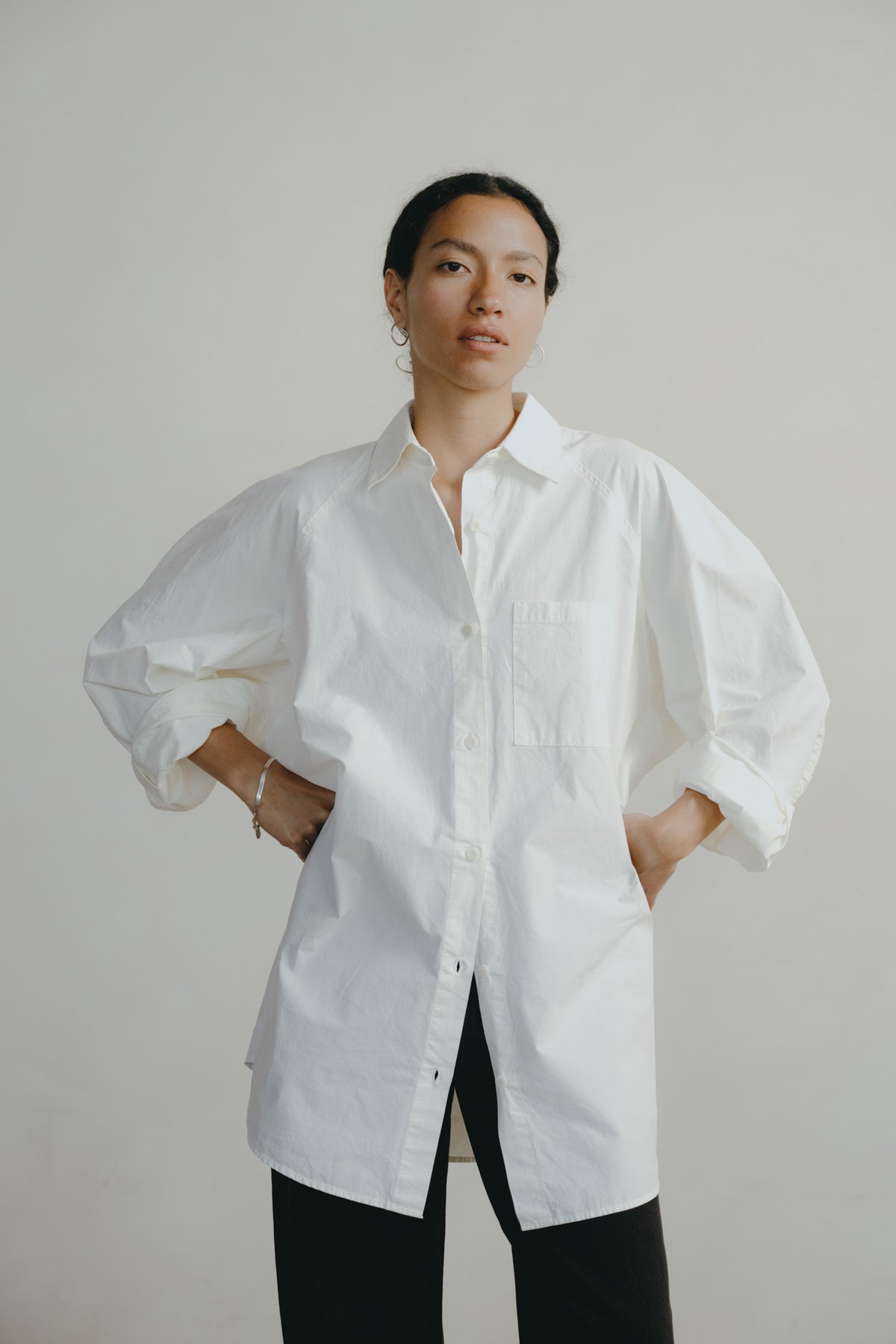 A woman modeling an oversized white button-up shirt with voluminous sleeves and a chest pocket, paired with black pants, against a plain light gray studio background.