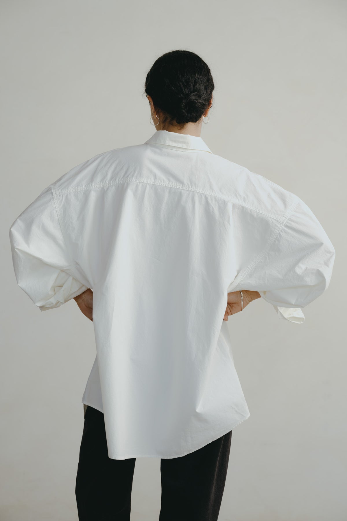 A person shown from the back wearing a loose white oversized button-down shirt with voluminous sleeves and black pants, standing against a plain light gray studio background.
