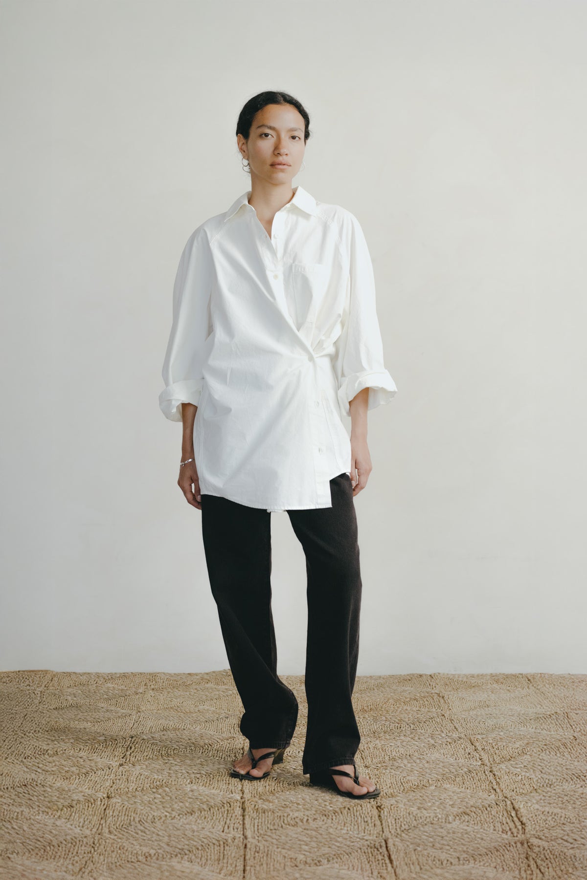 A model stands in a minimalist studio setting wearing a white oversized button-down shirt with rolled sleeves, black wide-leg trousers, and black sandals, standing on a textured beige woven rug against a plain off-white wall.