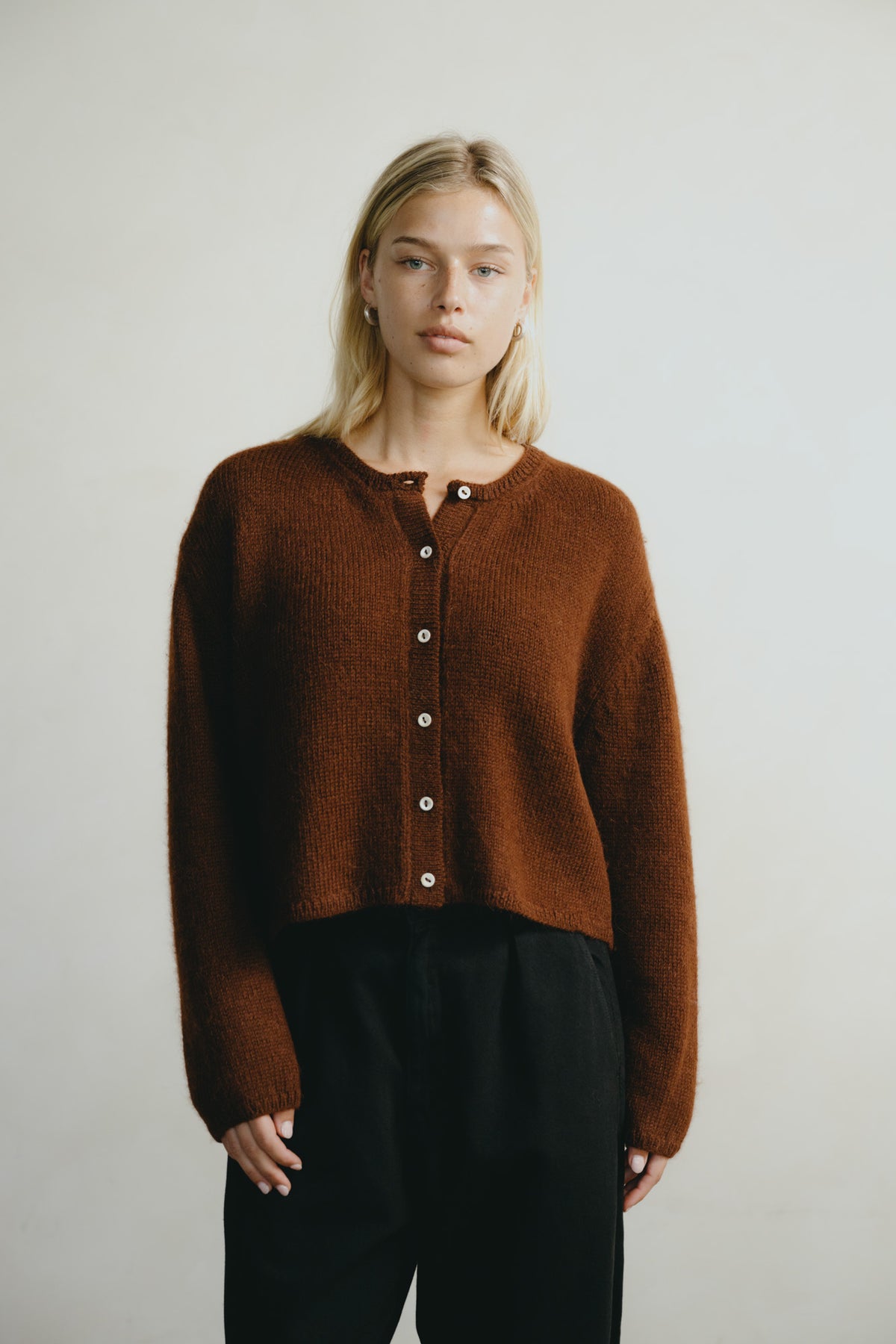 A blonde woman wearing a cropped, long-sleeve brown knit cardigan with white buttons and black trousers, standing against a plain light gray background.