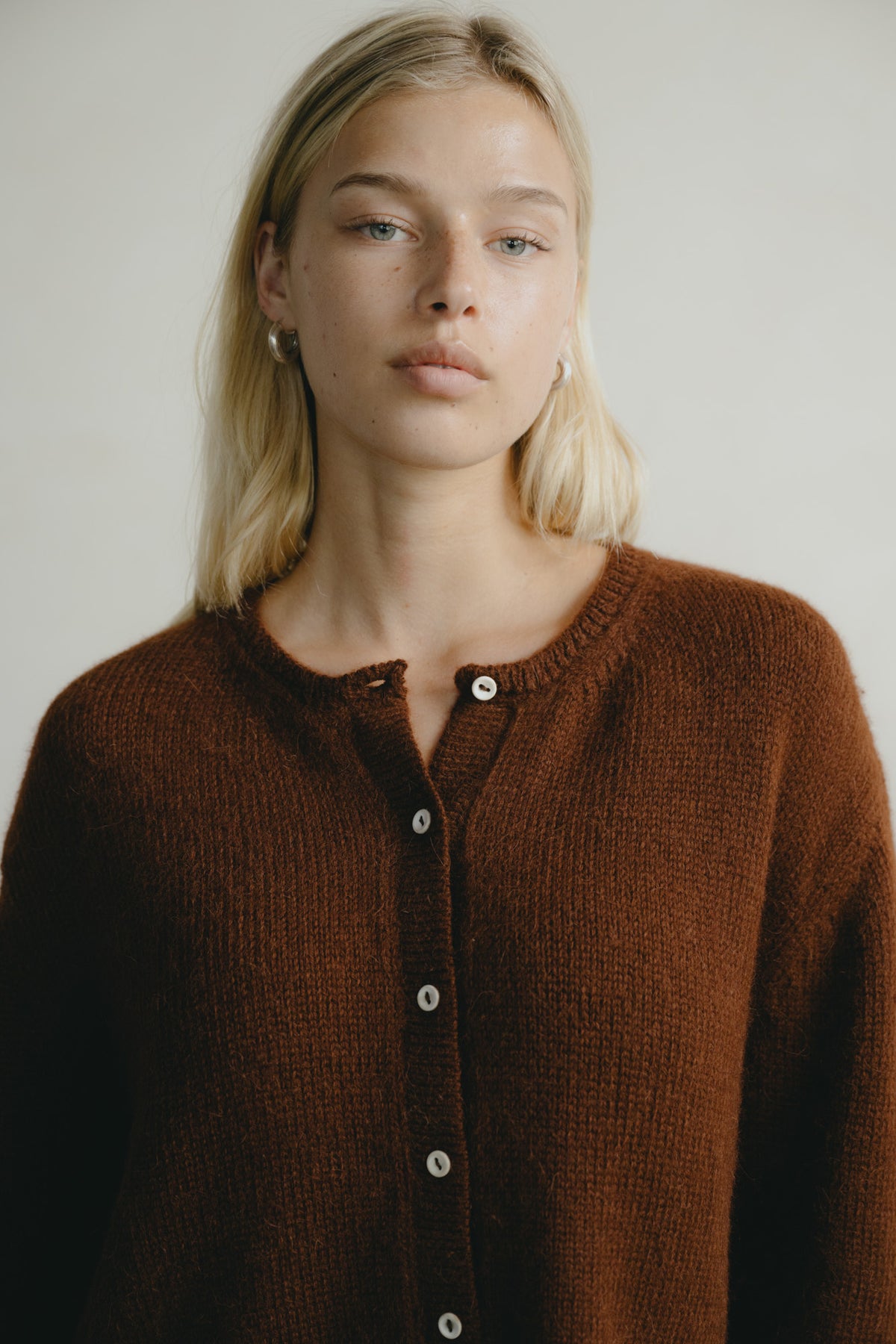 A blonde woman wearing a brown knit button-up cardigan sweater, photographed against a neutral light gray background.