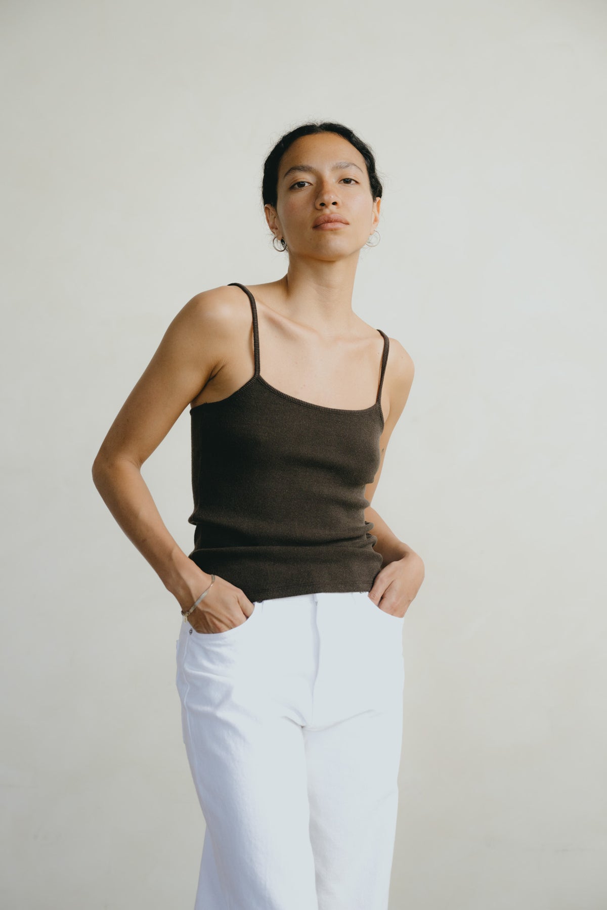 A woman modeling a dark brown ribbed knit camisole top with white pants, standing against a plain light gray background.