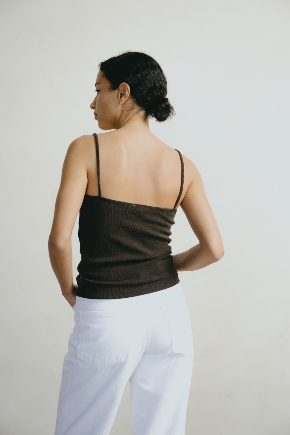A woman shown from the back wearing a dark brown spaghetti-strap camisole top and white pants, standing against a plain light gray studio background.