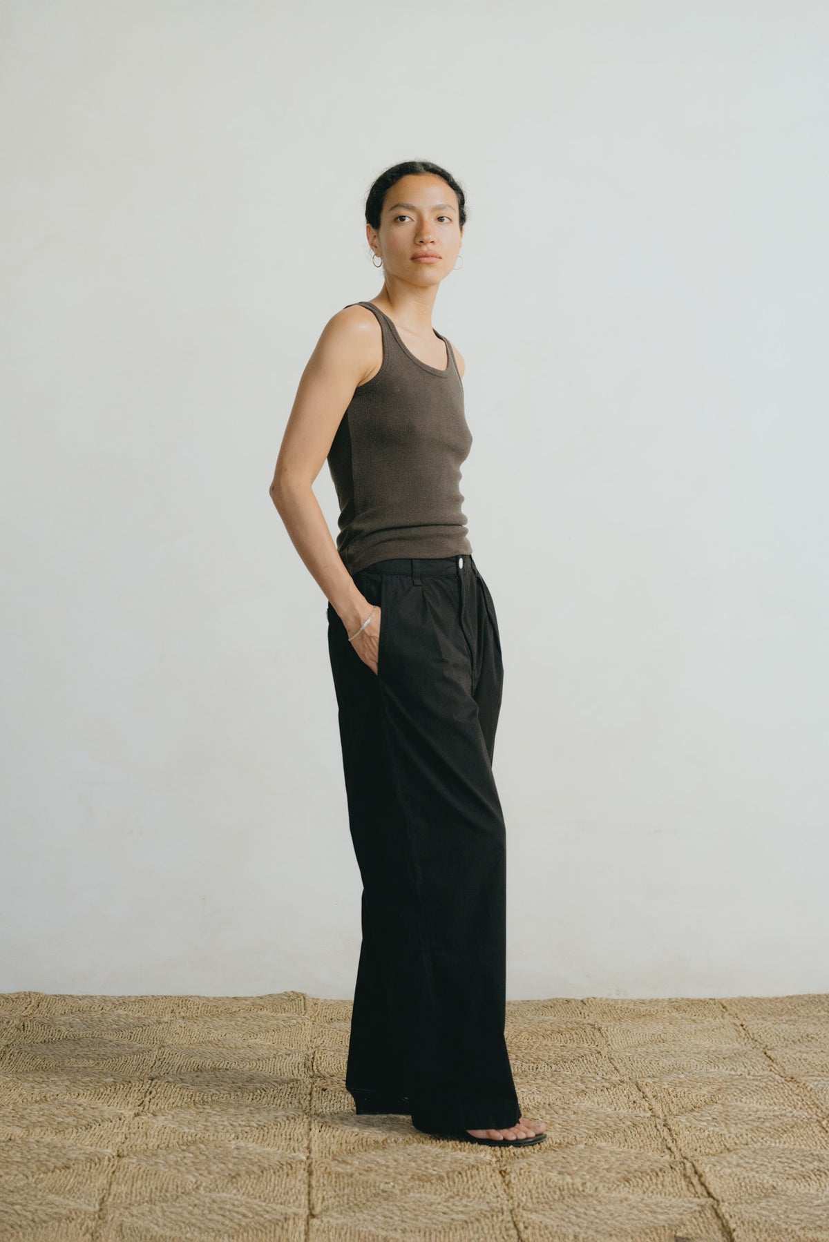 A woman stands in profile wearing a fitted taupe-brown ribbed tank top and loose black wide-leg pants, standing on a woven jute floor against a plain off-white wall.