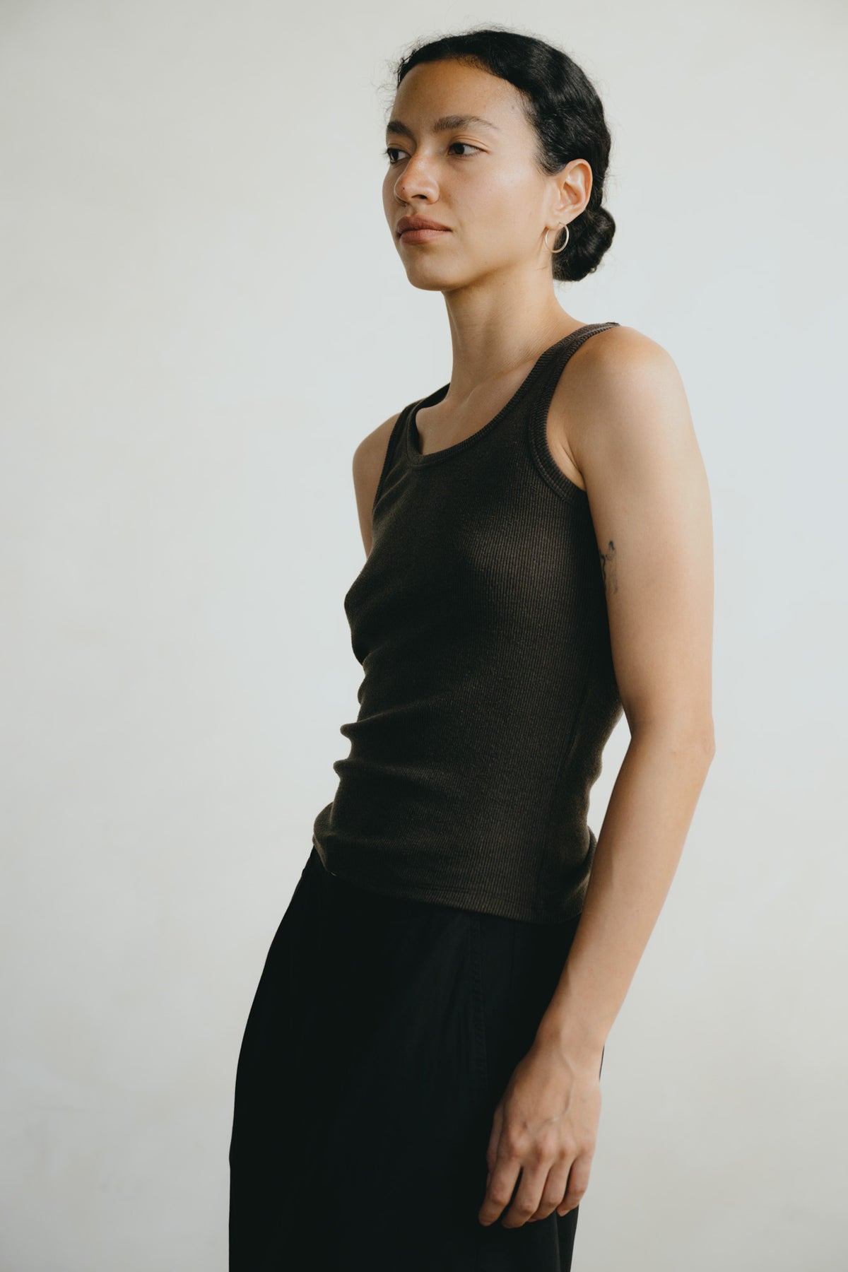 A woman wearing a dark brown ribbed tank top and black pants, standing in profile against a plain light gray background.