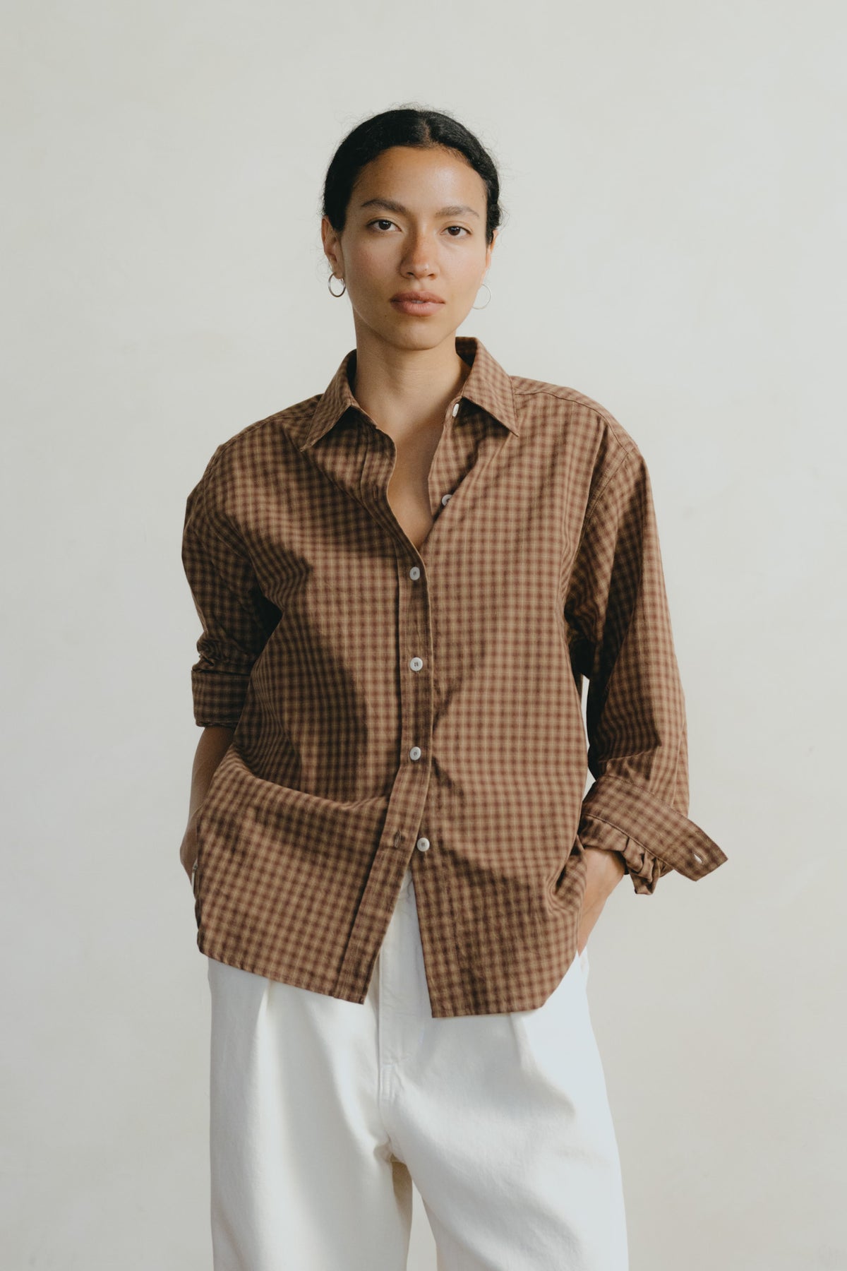 A woman wearing a brown and beige gingham button-up shirt with white trousers, standing against a plain light gray background.