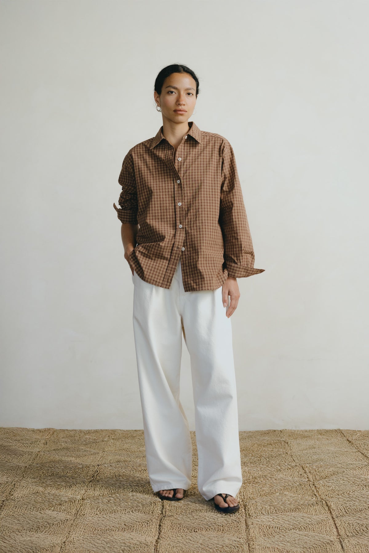 A model stands against a plain off-white wall wearing a brown and beige gingham button-up shirt and wide-leg white trousers, paired with dark sandals.