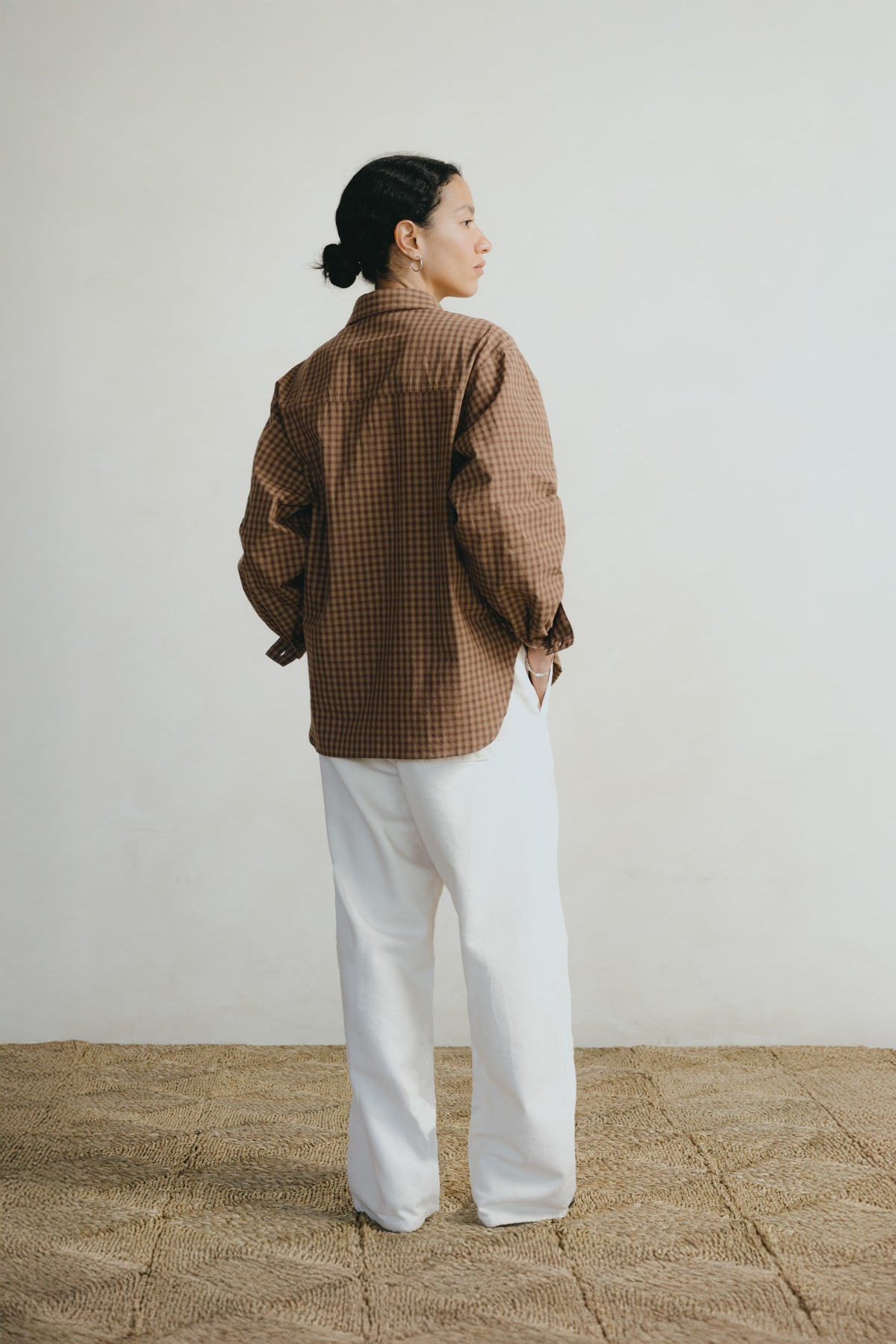 A woman stands with her back to the camera wearing a brown and black checkered long-sleeve shirt and white wide-leg pants, set against a plain off-white wall and a textured beige floor.