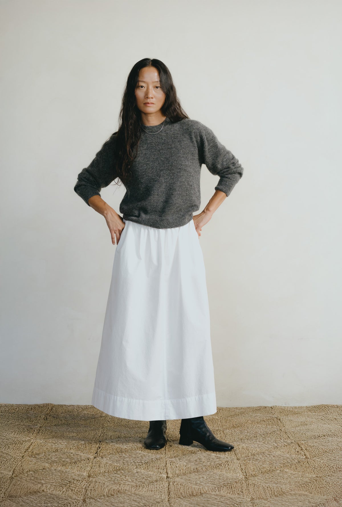 A woman stands with hands on hips wearing a dark gray knit sweater and a long white skirt, paired with black ankle boots, against a plain off-white wall and a woven jute floor.