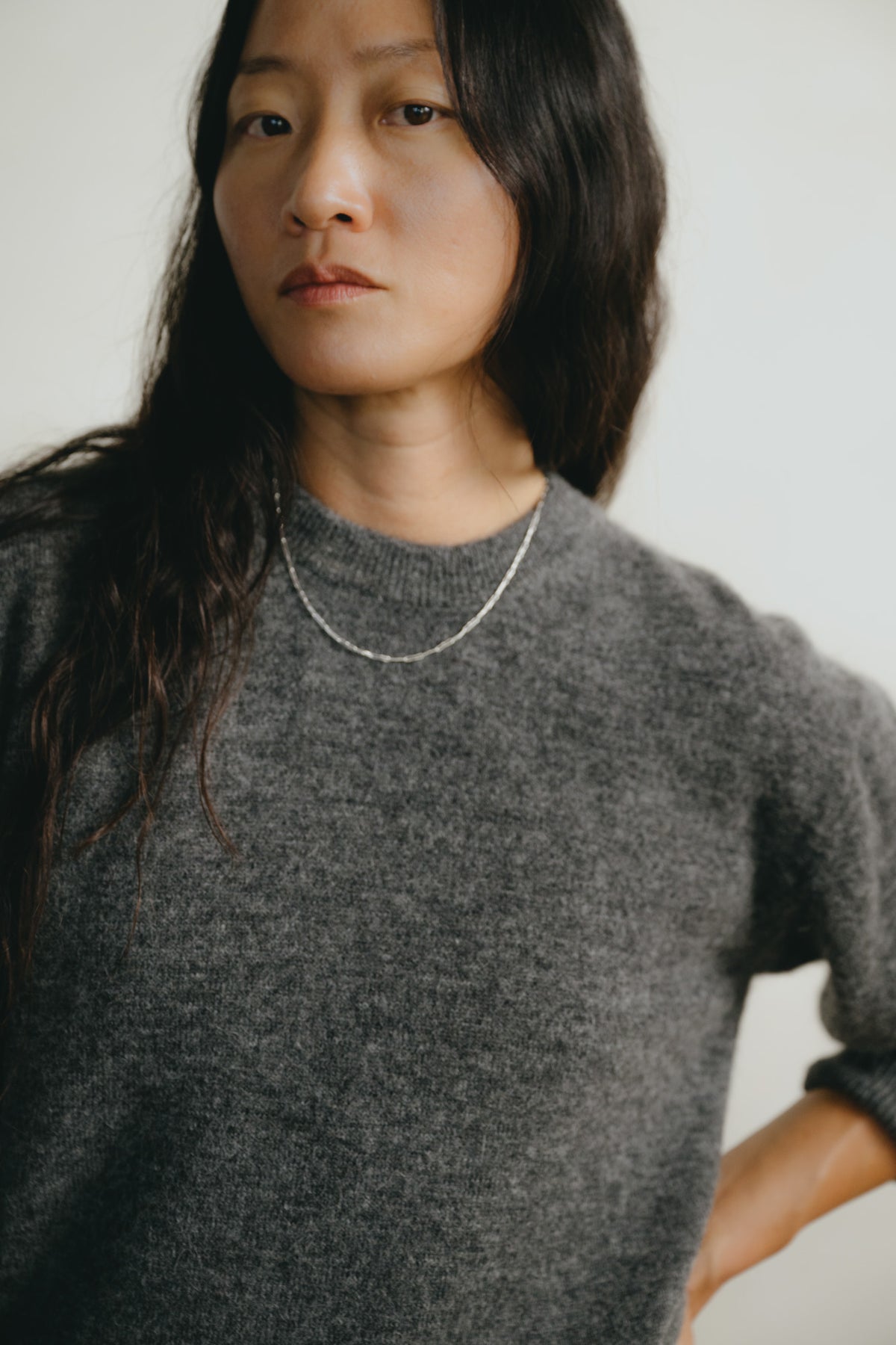 A close-up shot of a woman wearing a dark gray knit crewneck sweater and a delicate silver chain necklace, set against a plain light background.