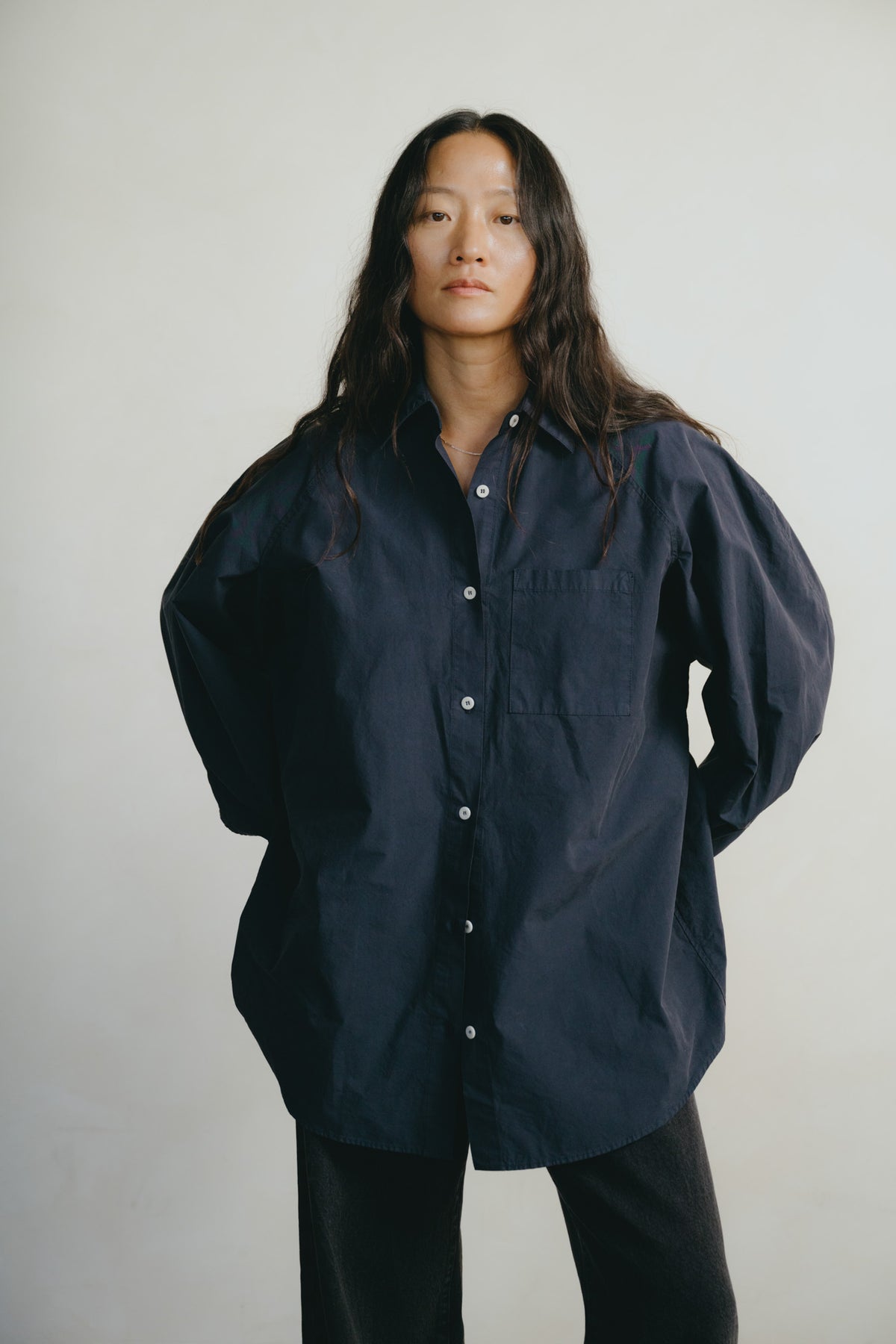 A woman wearing an oversized dark navy blue button-up shirt with voluminous long sleeves and black pants, standing against a plain light gray background.