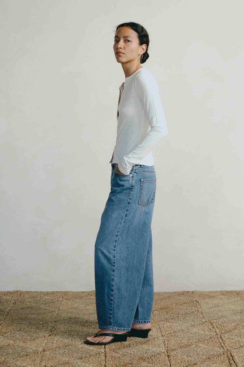 A woman stands in profile wearing a fitted white long-sleeve top, wide-leg blue jeans, and black sandals against a plain light wall and woven floor.