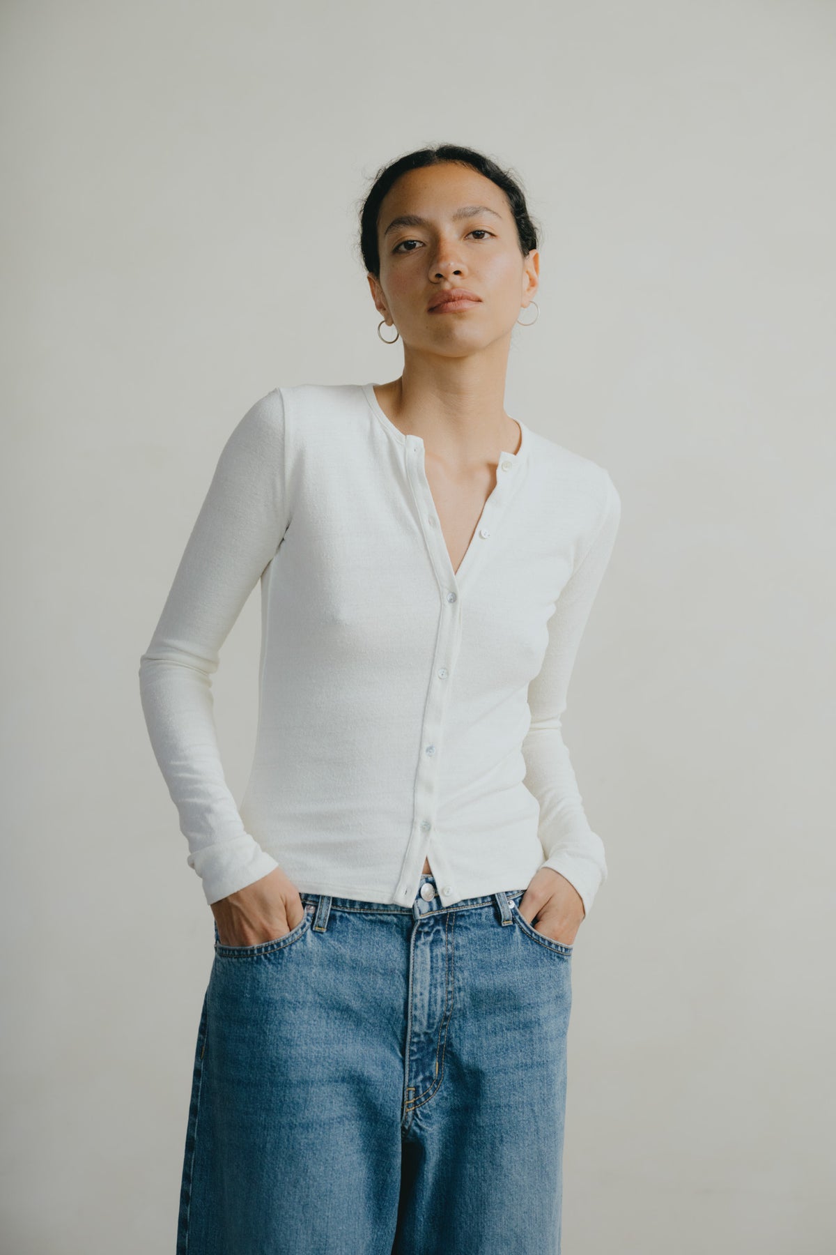 A woman wearing a fitted white long-sleeve button-front top and blue jeans, standing against a plain light gray studio background.