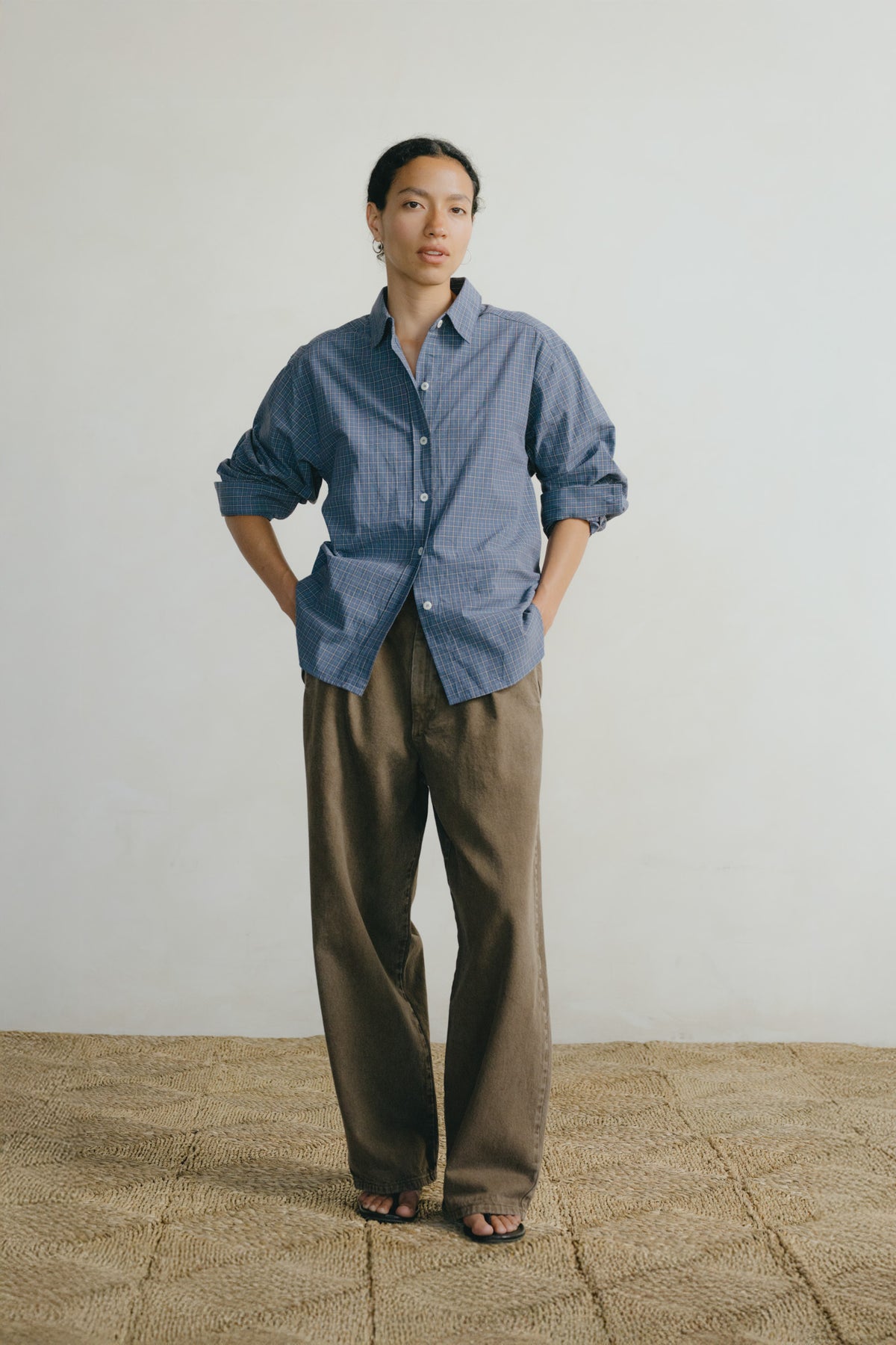 A model stands in a studio wearing a blue button-down shirt with rolled sleeves and wide-leg brown trousers, paired with black sandals, against a plain off-white wall and textured tan floor.