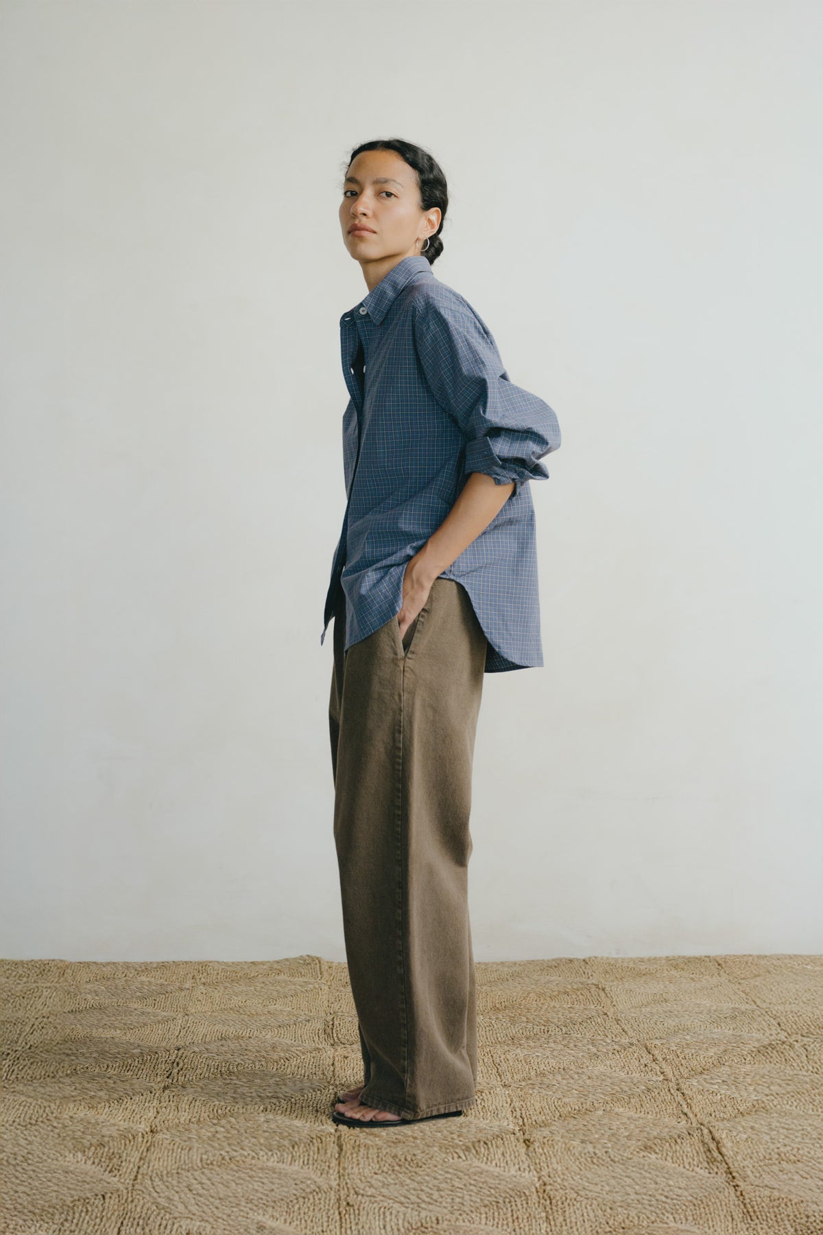 A model stands in profile wearing an oversized blue button-up shirt with rolled sleeves and brown wide-leg trousers, standing on a woven jute floor against a plain off-white wall.