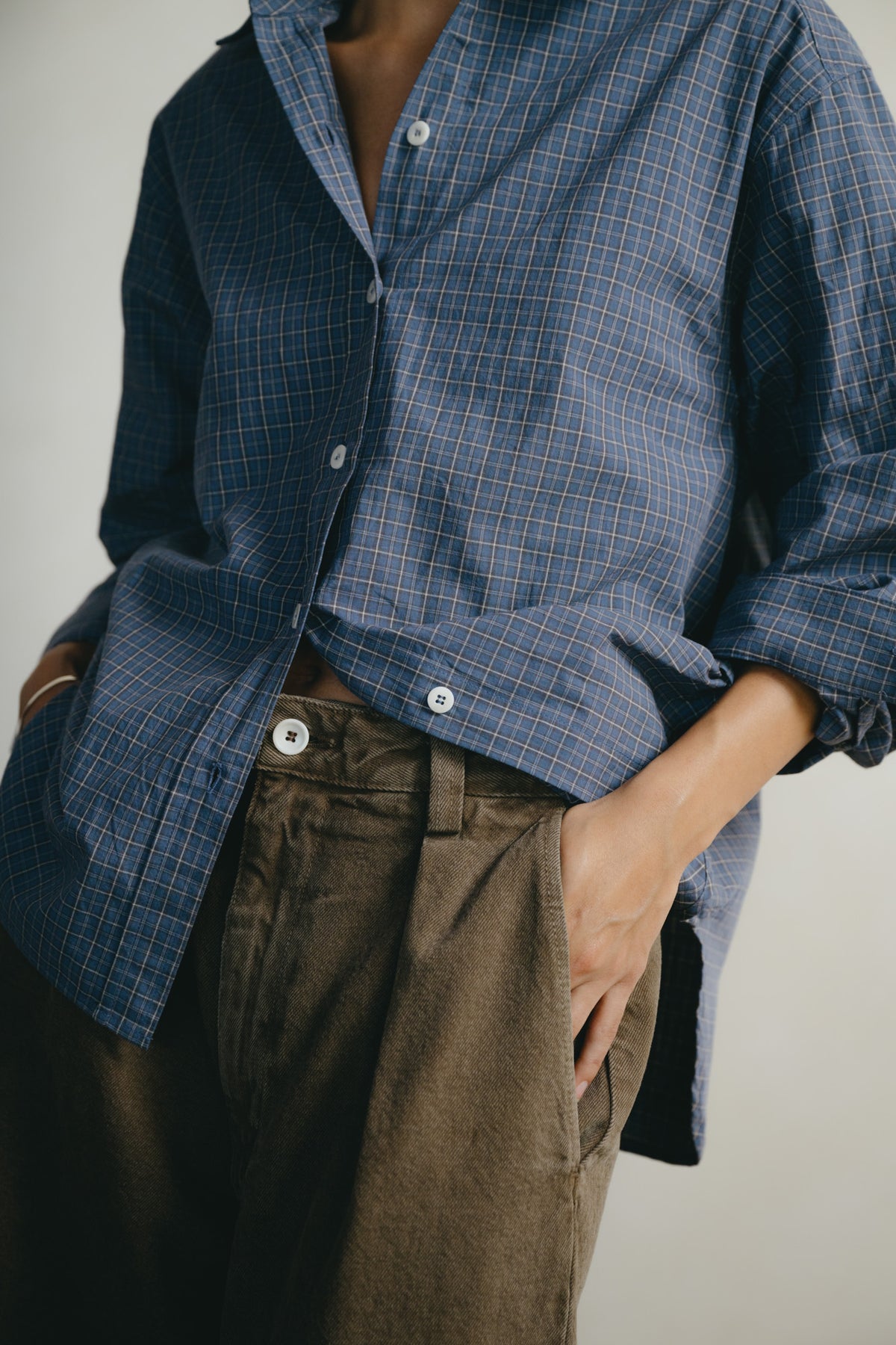 A person wearing a blue and gray plaid button-down shirt with rolled-up sleeves and brown trousers, shown from the neck down against a plain light background.