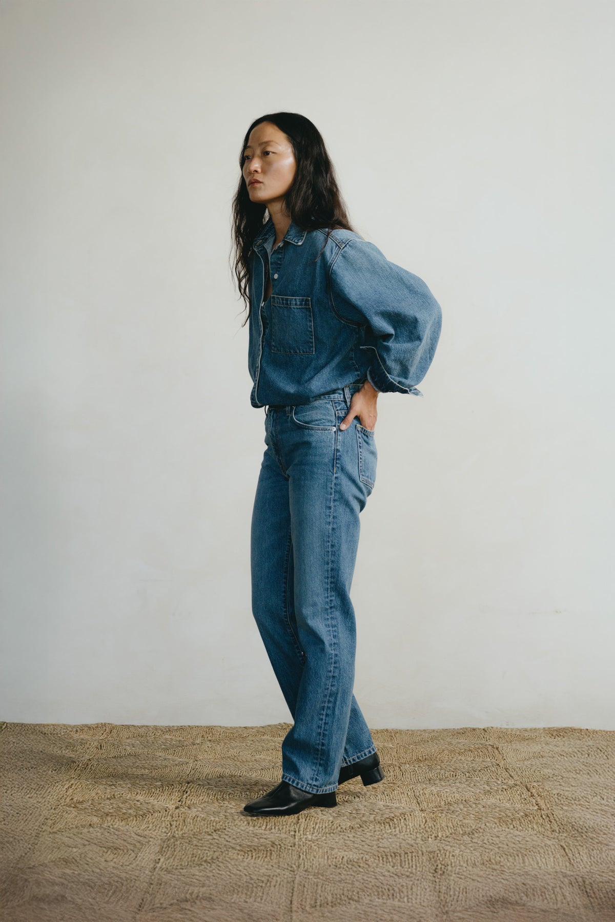 A woman stands in profile wearing a blue denim jacket with voluminous sleeves and matching blue denim jeans, paired with black shoes, against a plain off-white wall and textured tan floor.