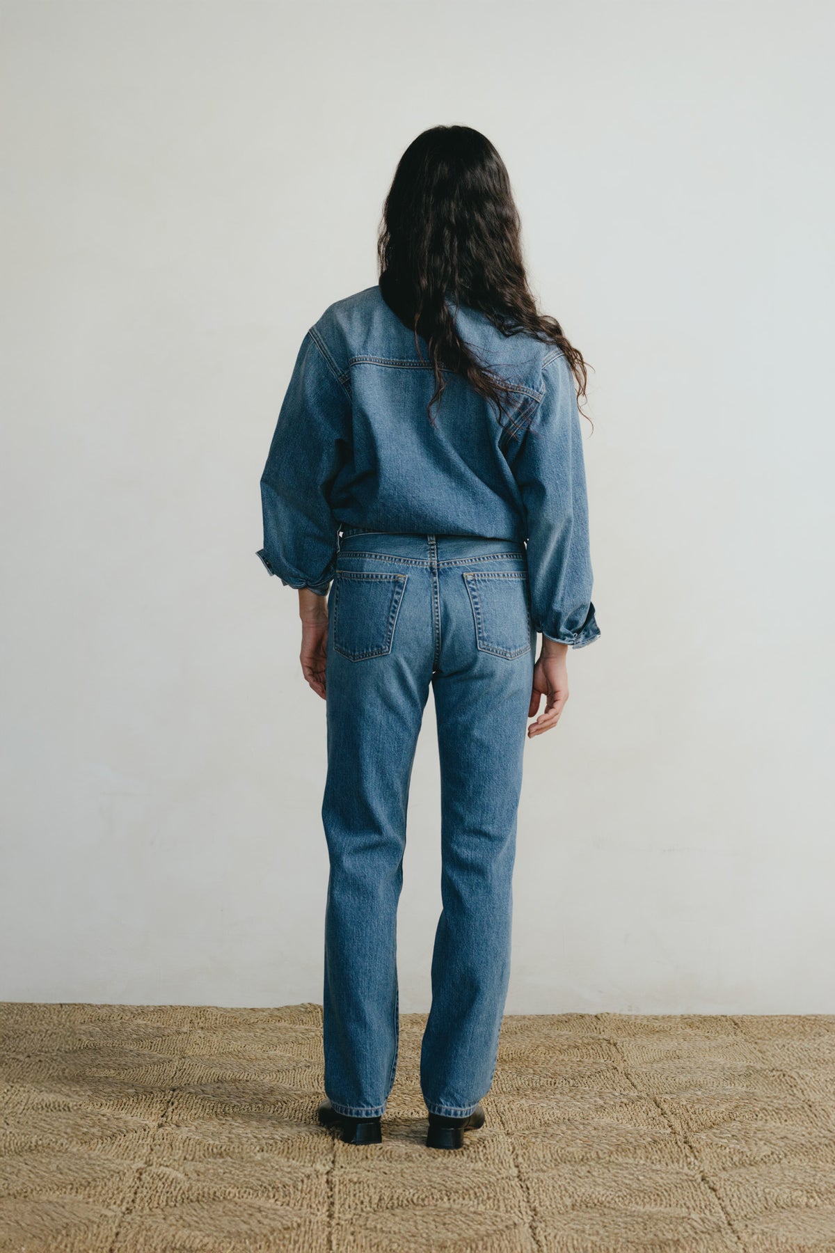 A woman viewed from behind wearing a blue denim shirt and matching blue denim jeans, standing on a woven rug against a plain off-white wall.