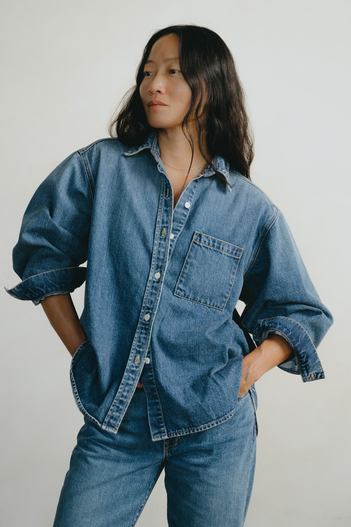A woman wearing an oversized blue denim button-up shirt with rolled sleeves and matching blue jeans, standing against a plain light gray background.