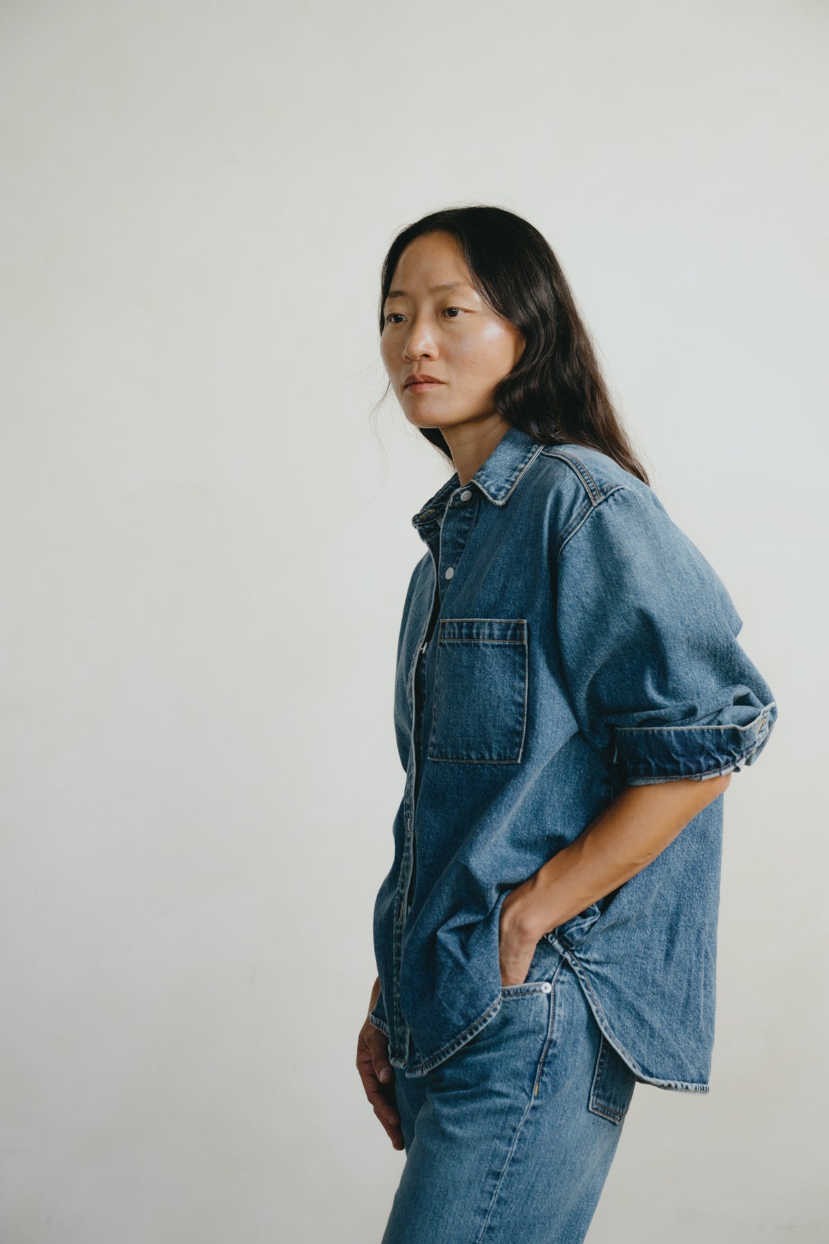 A woman wearing an oversized blue denim shirt with rolled sleeves and matching blue denim pants, standing against a plain light gray studio background.
