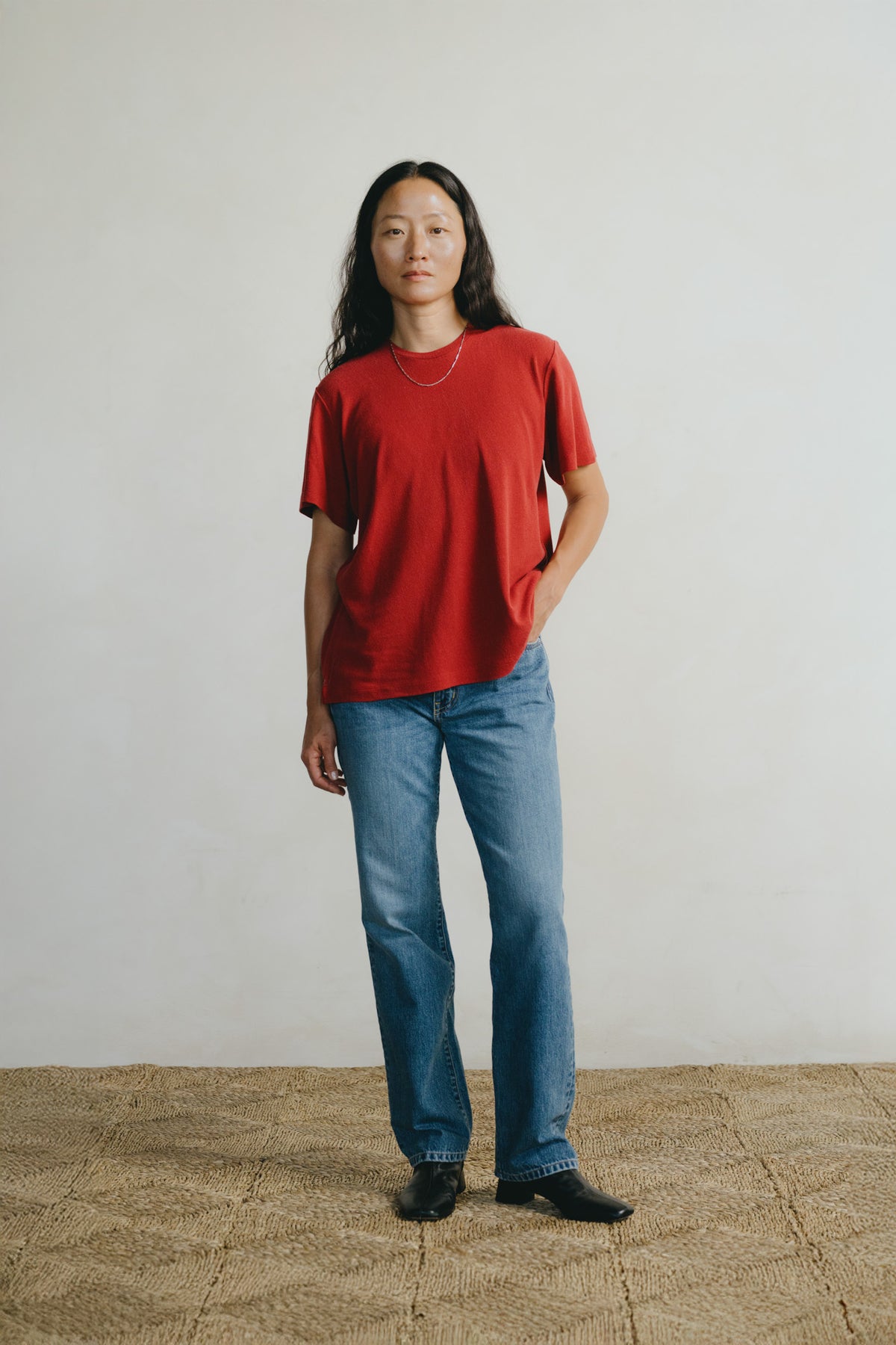 A woman stands in a studio wearing a loose red short-sleeve T-shirt, blue jeans, and black shoes against a plain light wall and woven floor.
