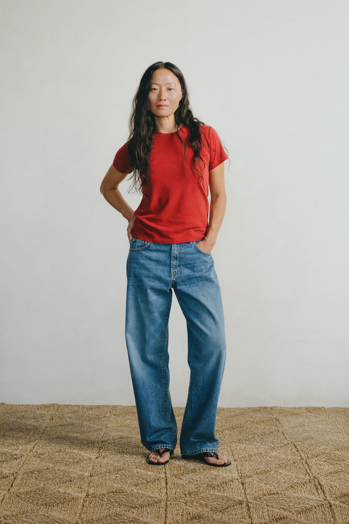 A woman stands in a studio wearing a red short-sleeve crewneck t-shirt, loose-fit blue jeans, and dark sandals. She is posed against a plain off-white wall with a textured tan woven rug on the floor.