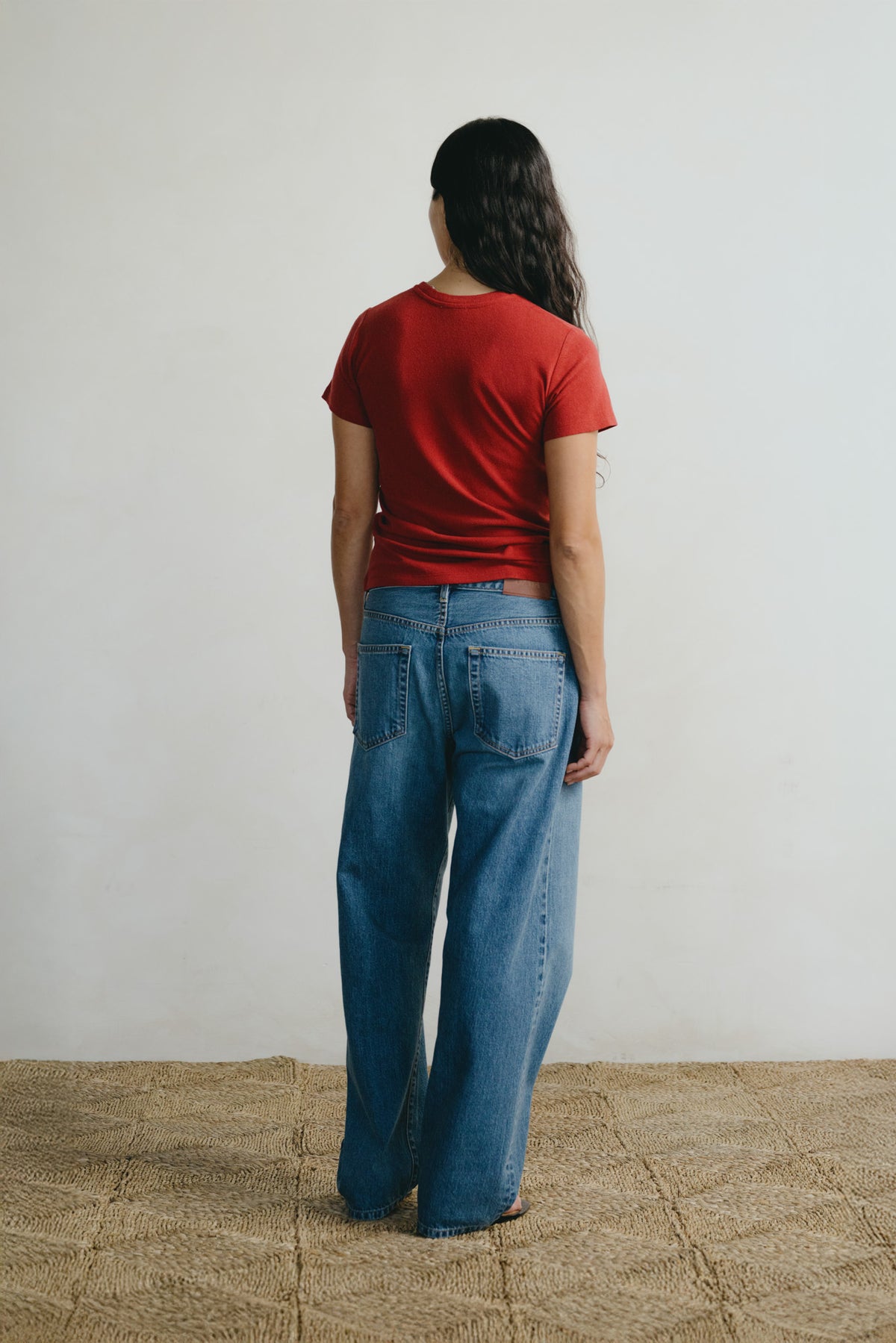 A woman viewed from behind wearing a red short-sleeve t-shirt and wide-leg blue jeans, standing on a textured tan floor against a plain off-white wall.