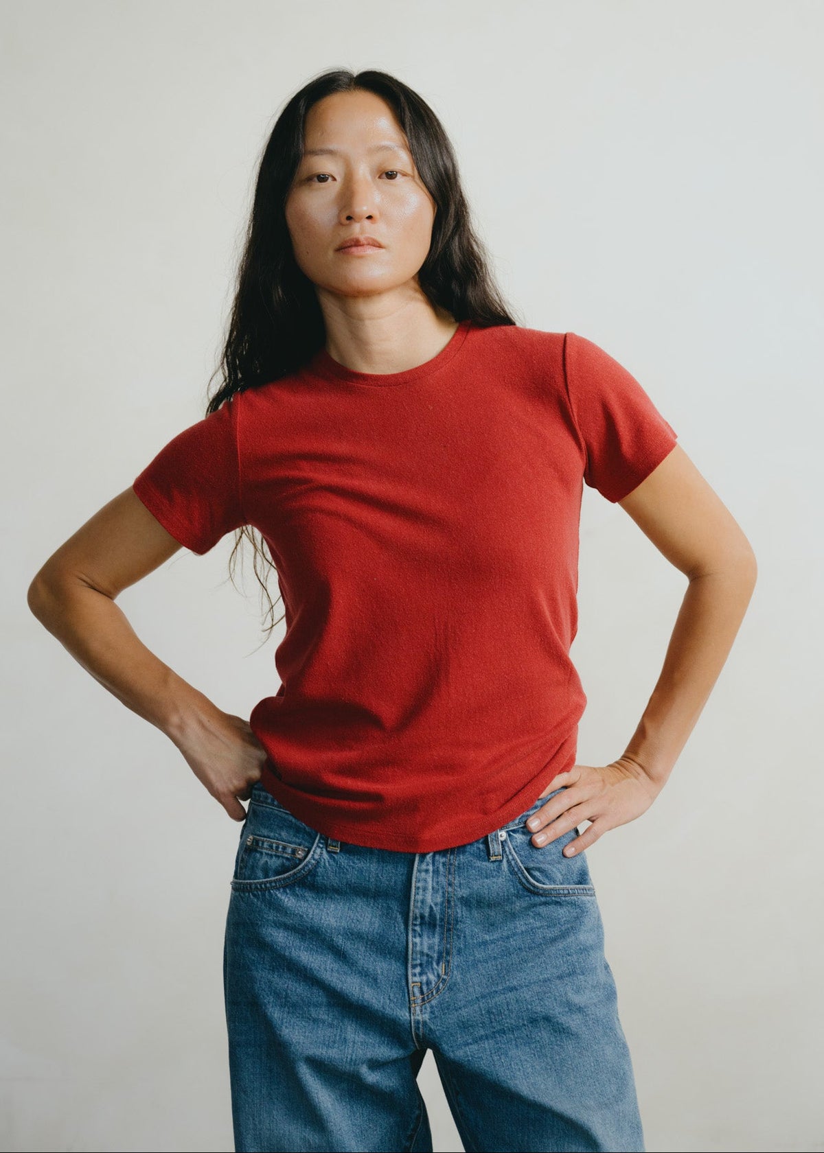 A woman with long dark hair wearing a solid red short-sleeve crewneck t-shirt and blue jeans, standing against a plain light gray background with her hands on her hips.