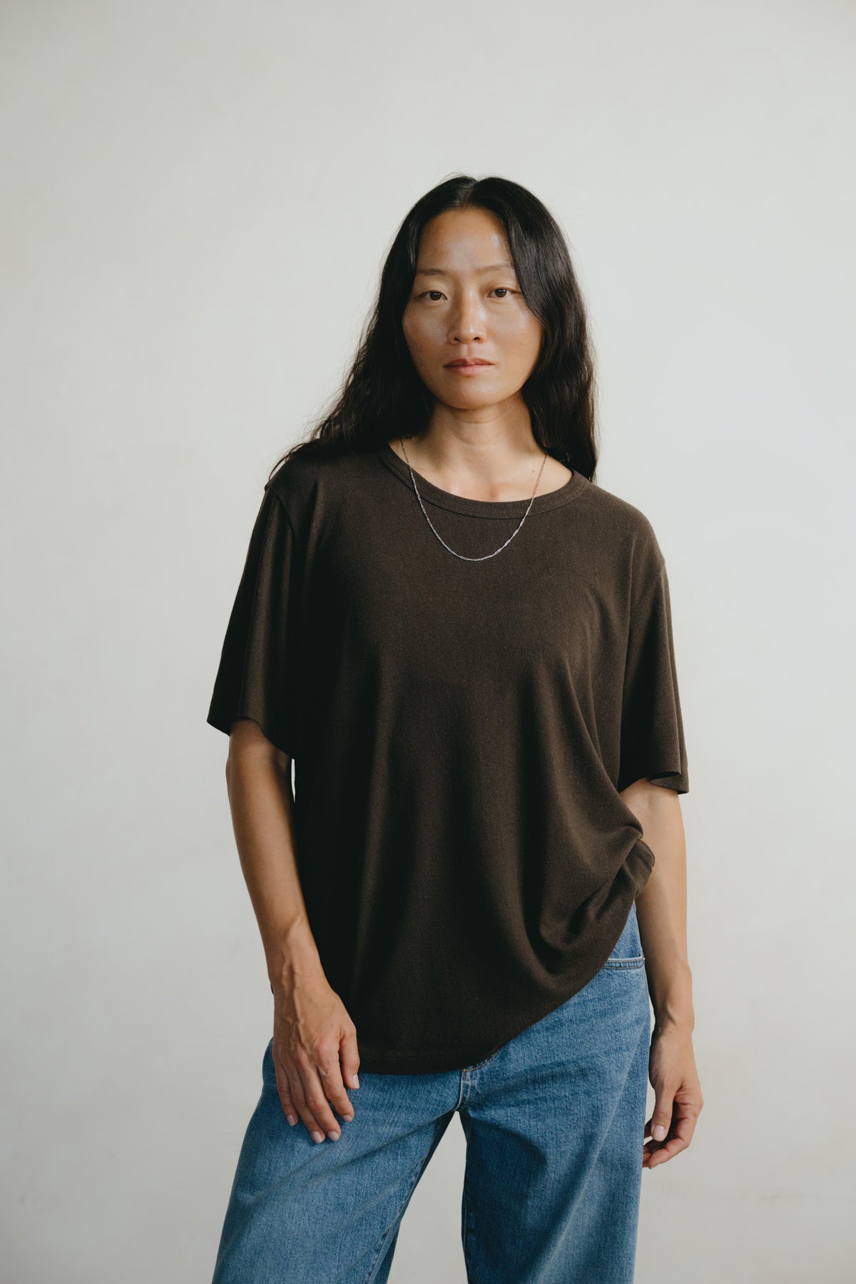 A woman wearing a dark brown oversized short-sleeve t-shirt and blue jeans, standing against a plain light gray background.