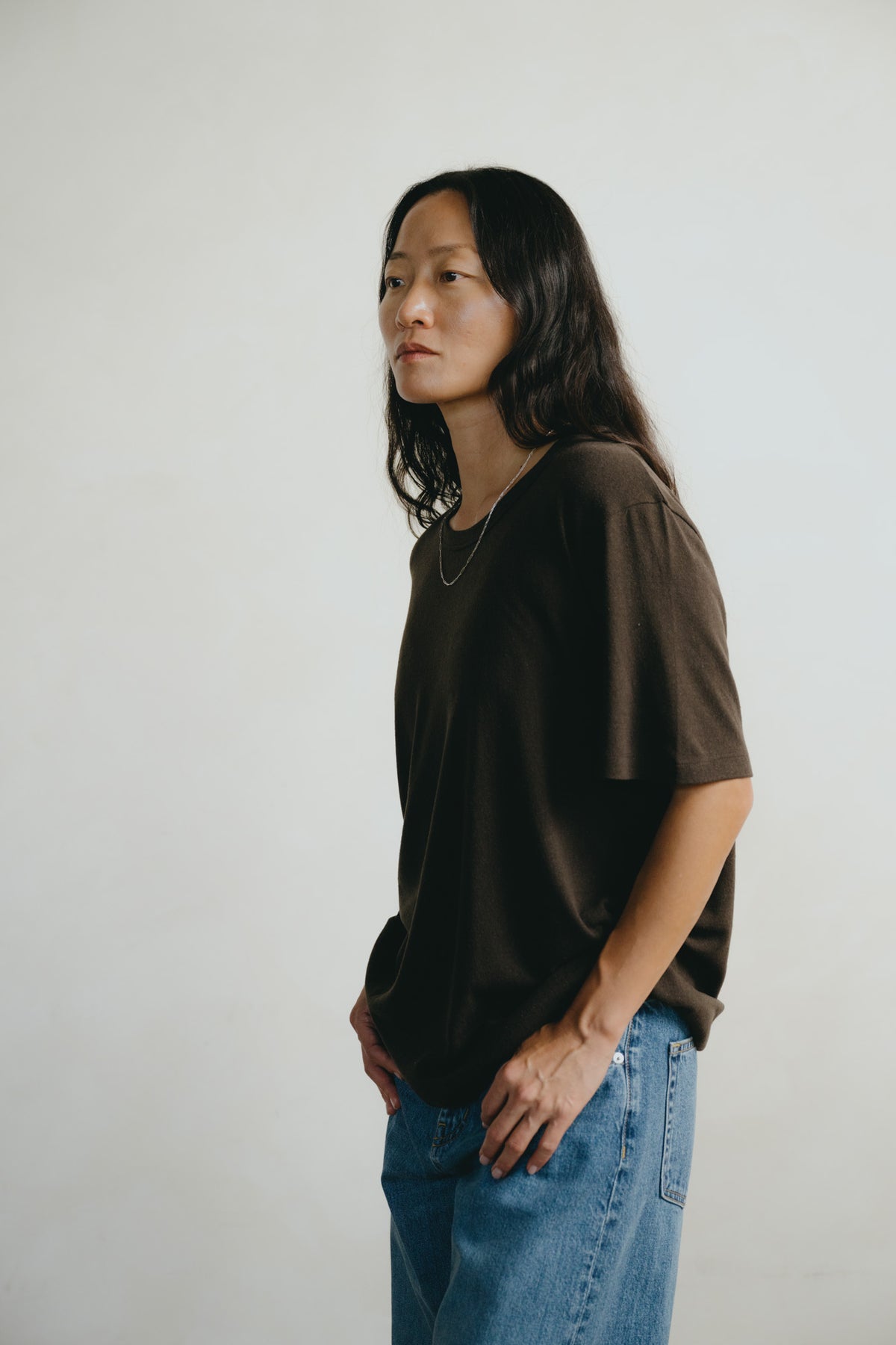 A woman with long dark hair is shown in profile wearing an oversized dark brown t-shirt and blue jeans against a plain light gray background.